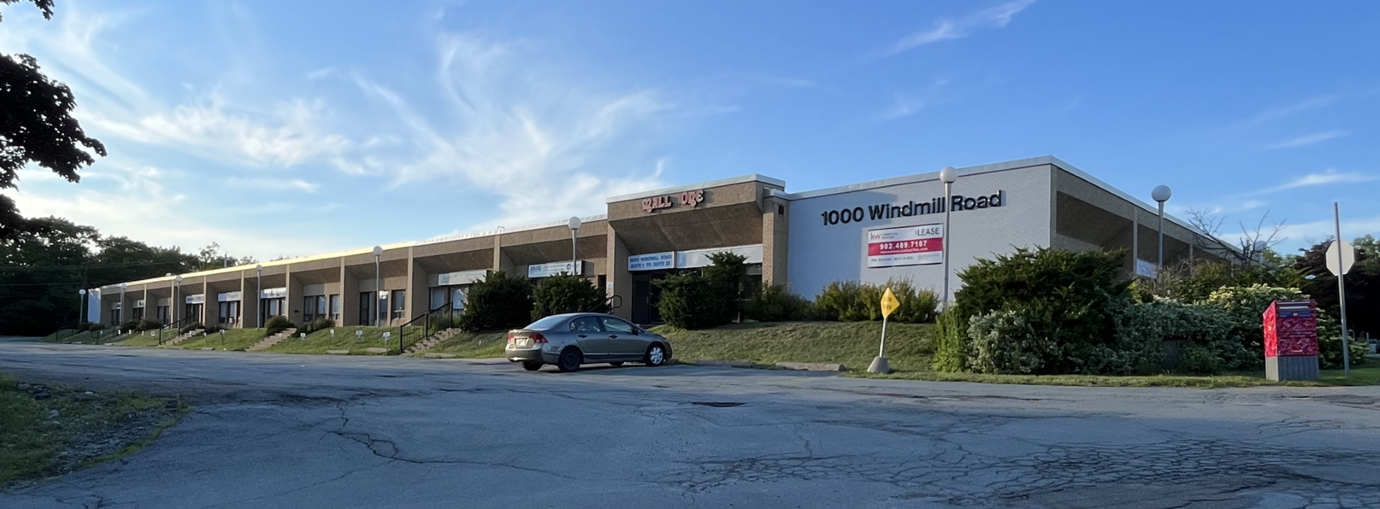 A single-story commercial building with multiple units at 1000 Windmill Road, with a parking lot in front. The building has a sign "Wall One" and some lease advertisements, and is surrounded by bushes and trees, under a clear blue sky.