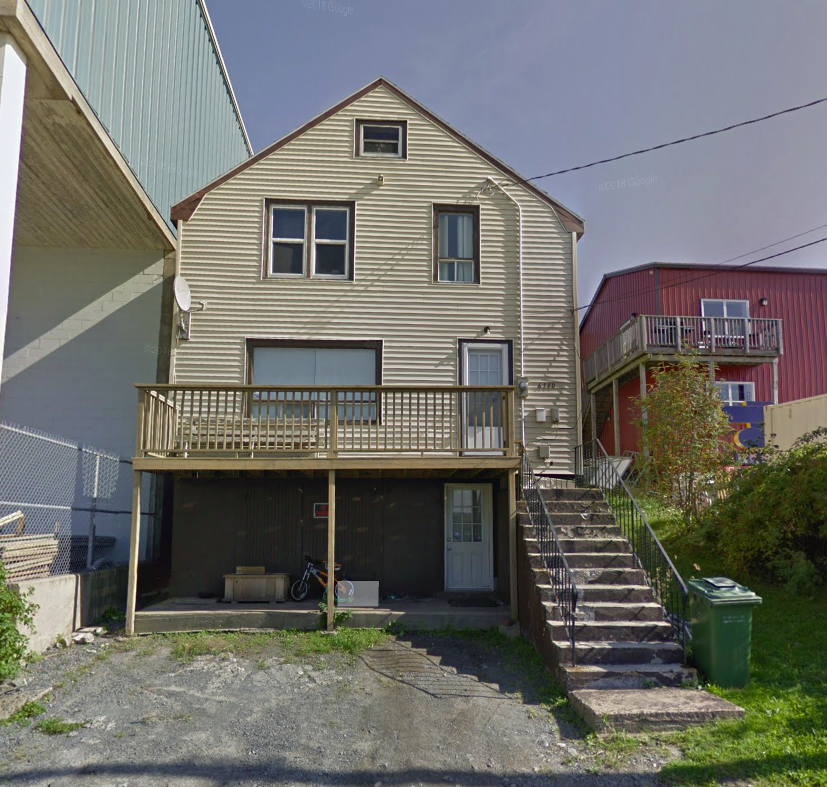 A multi-story house with a beige exterior and a wooden balcony on the second floor. The house has a staircase leading to the main door on the right side and a garage door on the lower level. There are neighboring houses visible on either side, one blue and one red.