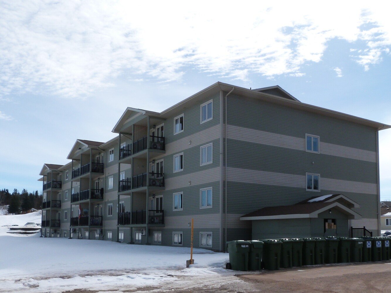 Multi-story residential apartment building with balconies, snow on the ground, and several trash bins outside.