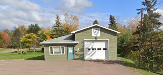 A green garage with a white roll-up door and a small door to the left, situated on a paved driveway with trees and colorful foliage in the background.