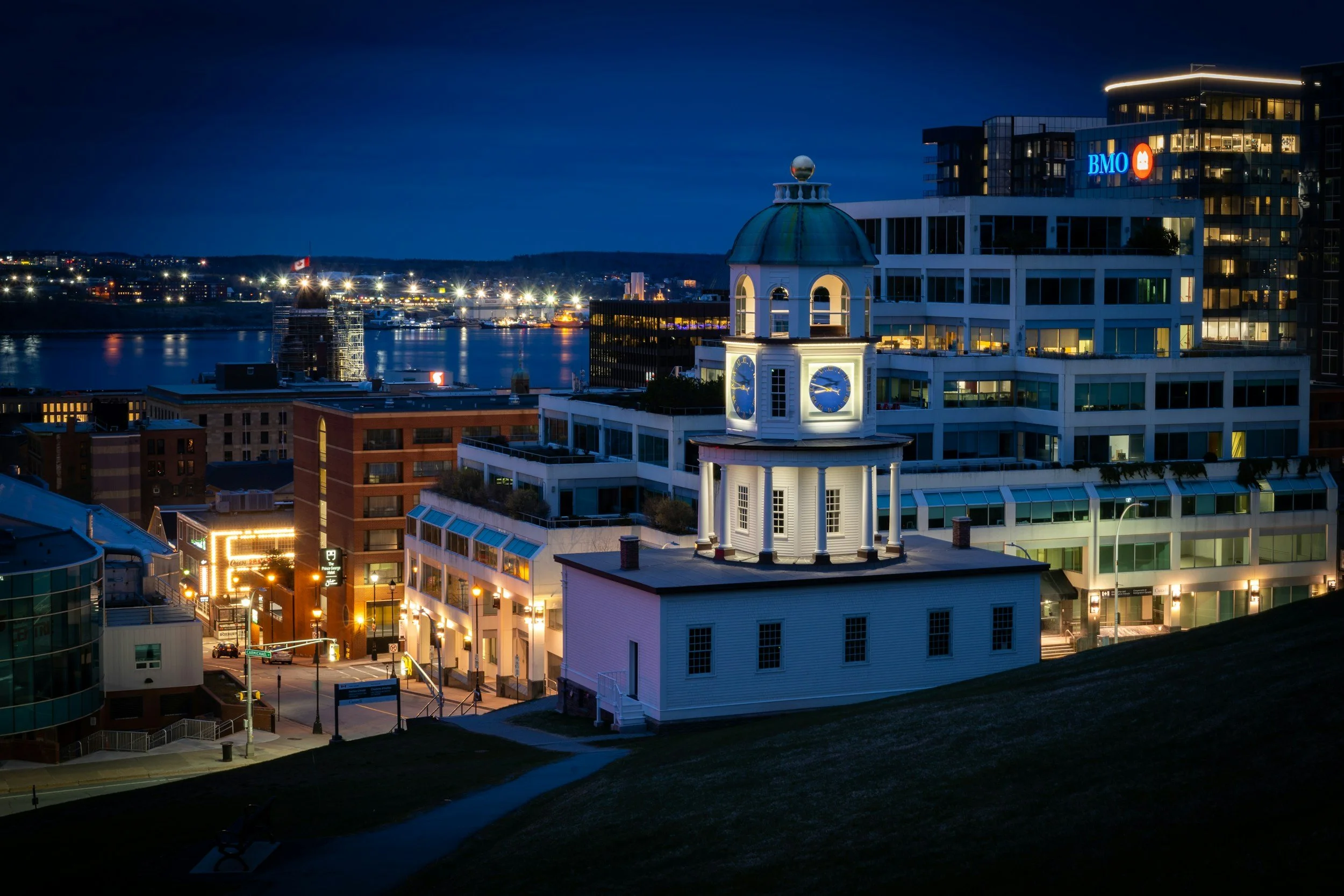 Nighttime cityscape with illuminated clock tower in foreground, modern glass buildings, and a river reflecting city lights, with a BMO bank sign on a building in the background.