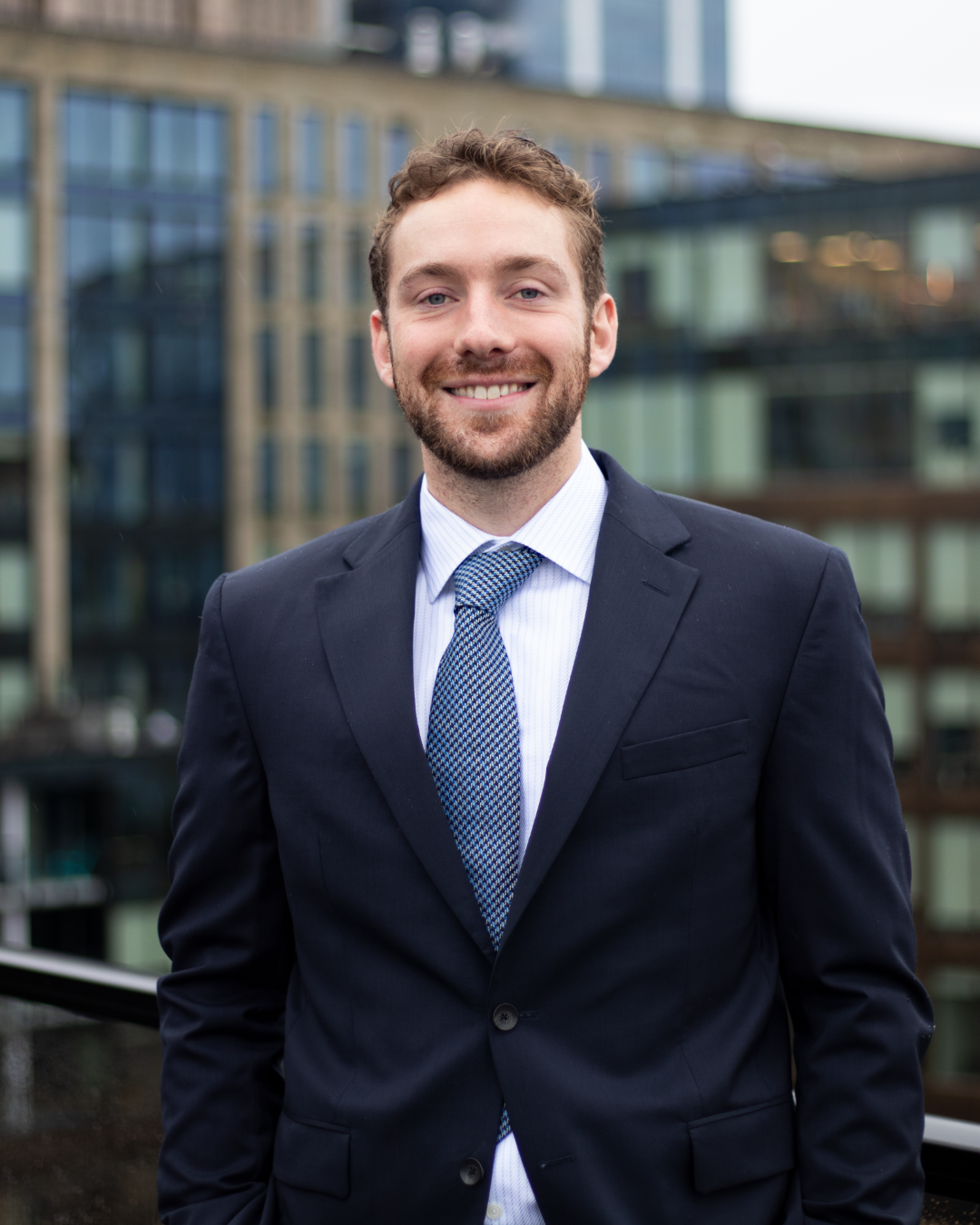 Headshot of a young man with brown hair and a beard, wearing a navy suit jacket, light blue shirt, and patterned tie, standing outdoors in front of modern glass buildings.