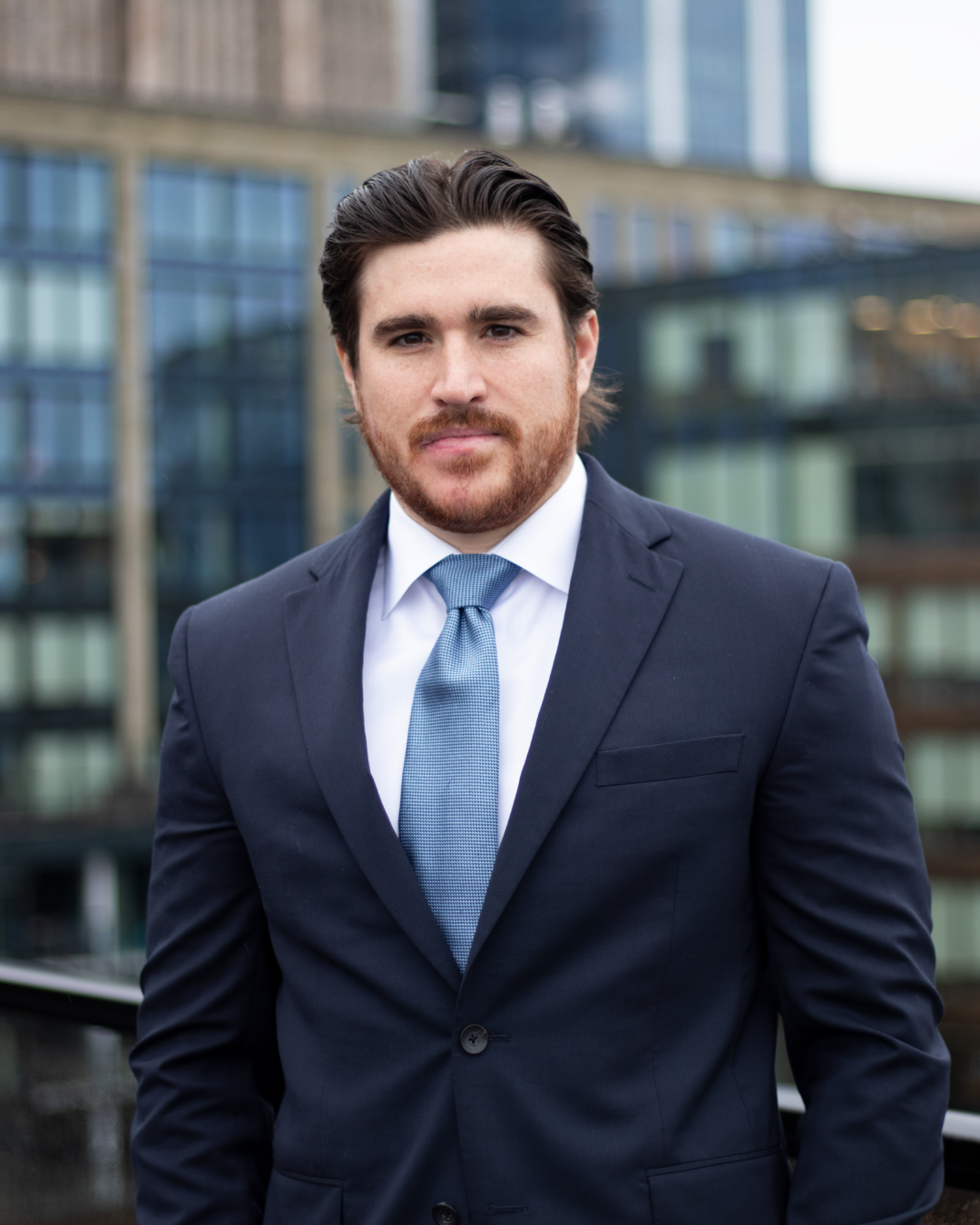 A man in a dark suit with a light blue tie, standing outdoors in front of a modern glass building.