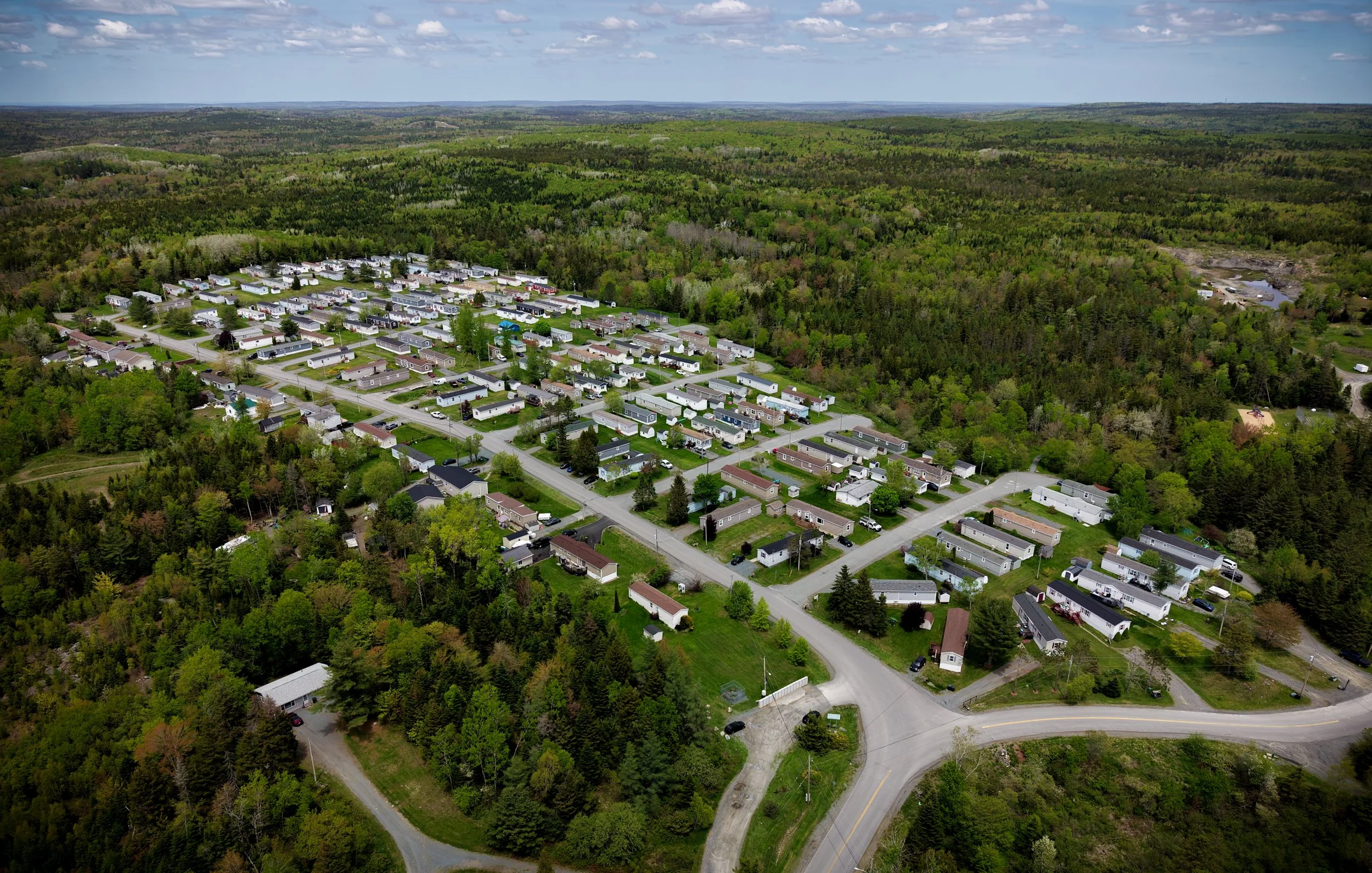 An aerial view of a mobile home community surrounded by dense green forests and rolling hills under a partly cloudy sky.