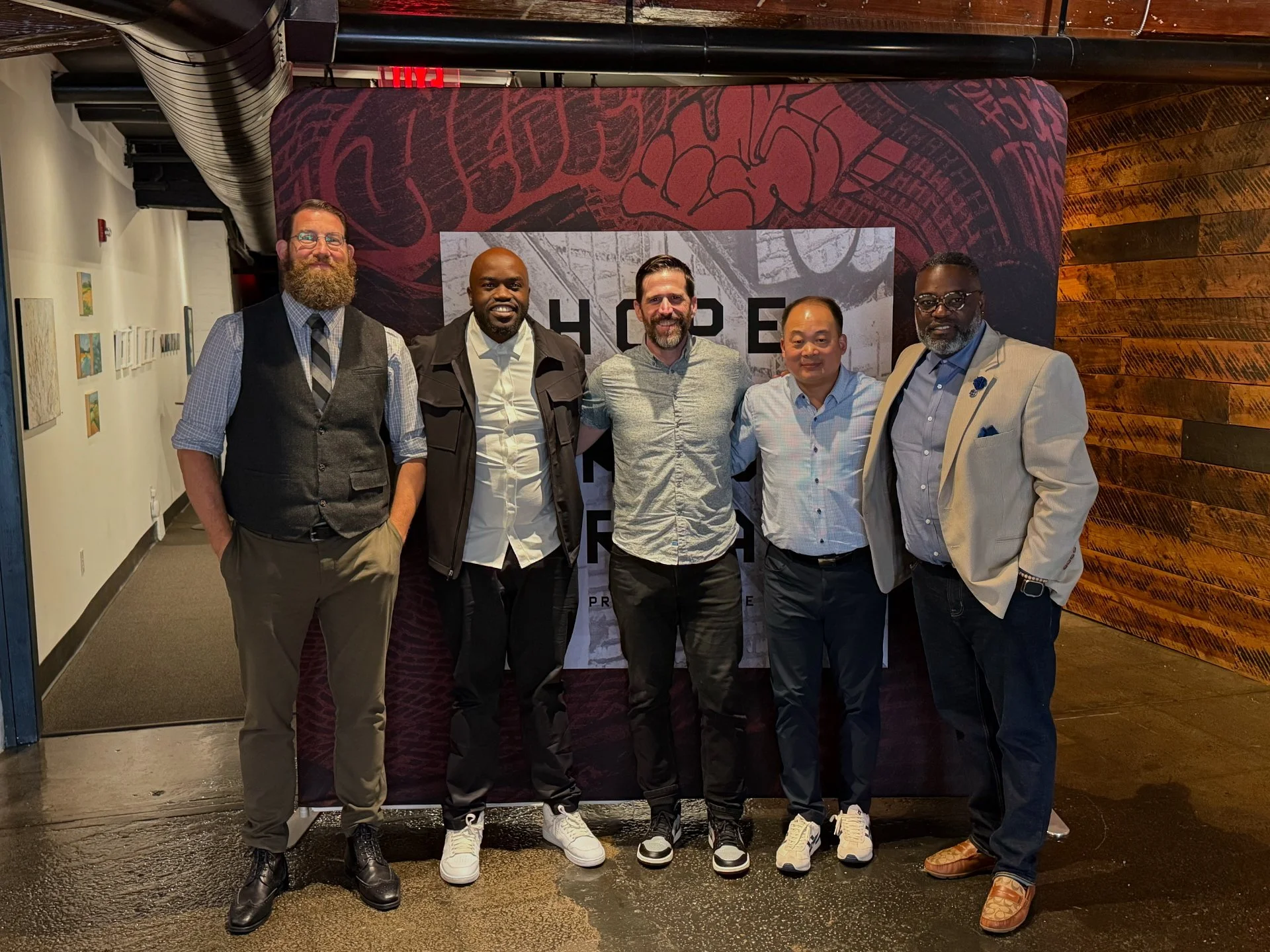 Five men standing together in front of a backdrop that says 'SHOE F R'. The men are dressed in business casual to formal attire, and they are indoors in what appears to be a gallery or event space with artwork on the wall.