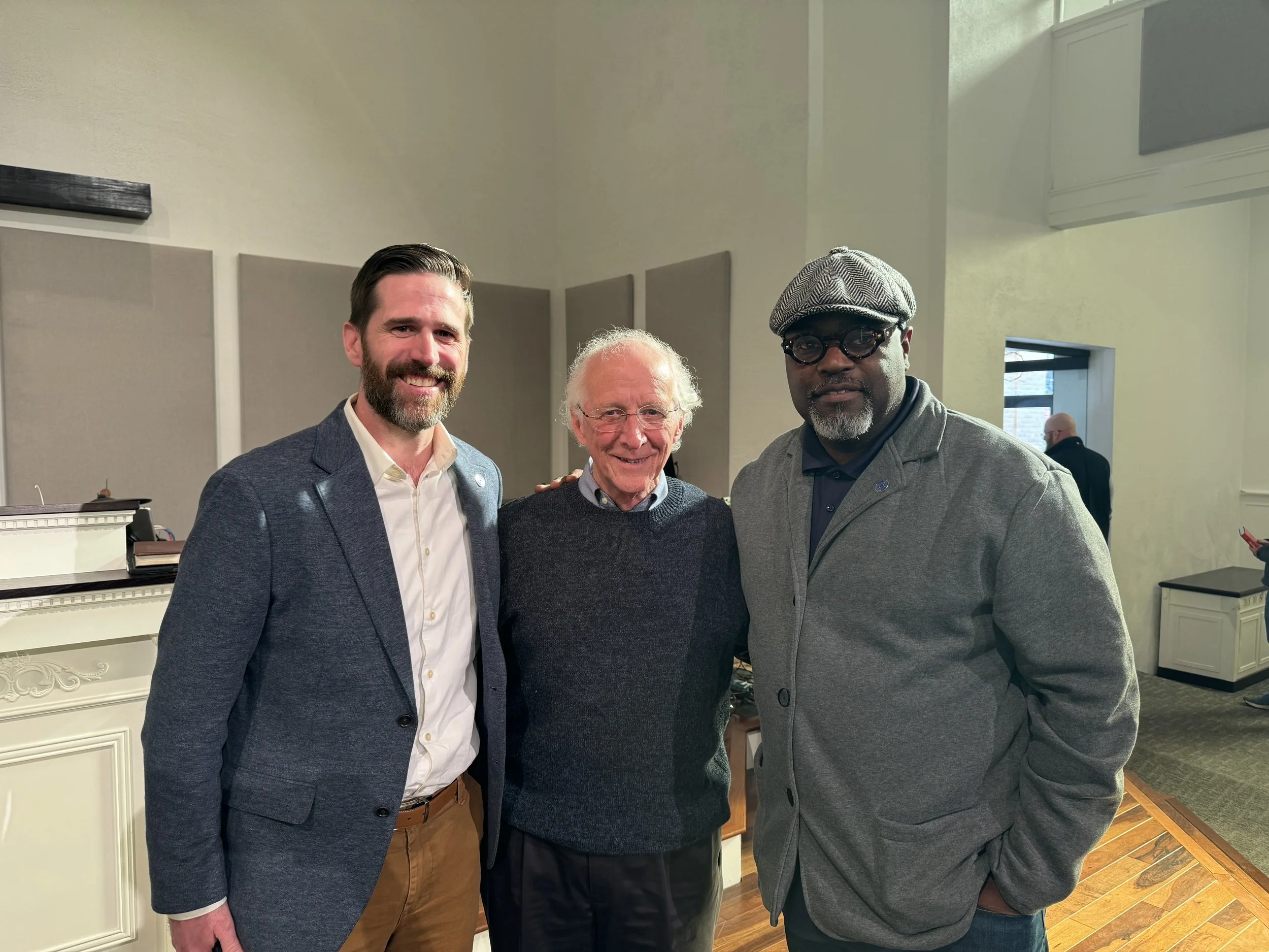 Three men standing together indoors, smiling for the photo. The man on the left has a beard and is dressed in a dark blue blazer and white shirt. The man in the middle has white hair and glasses, wearing a dark sweater. The man on the right is wearing glasses, a gray flat cap, and a gray jacket.