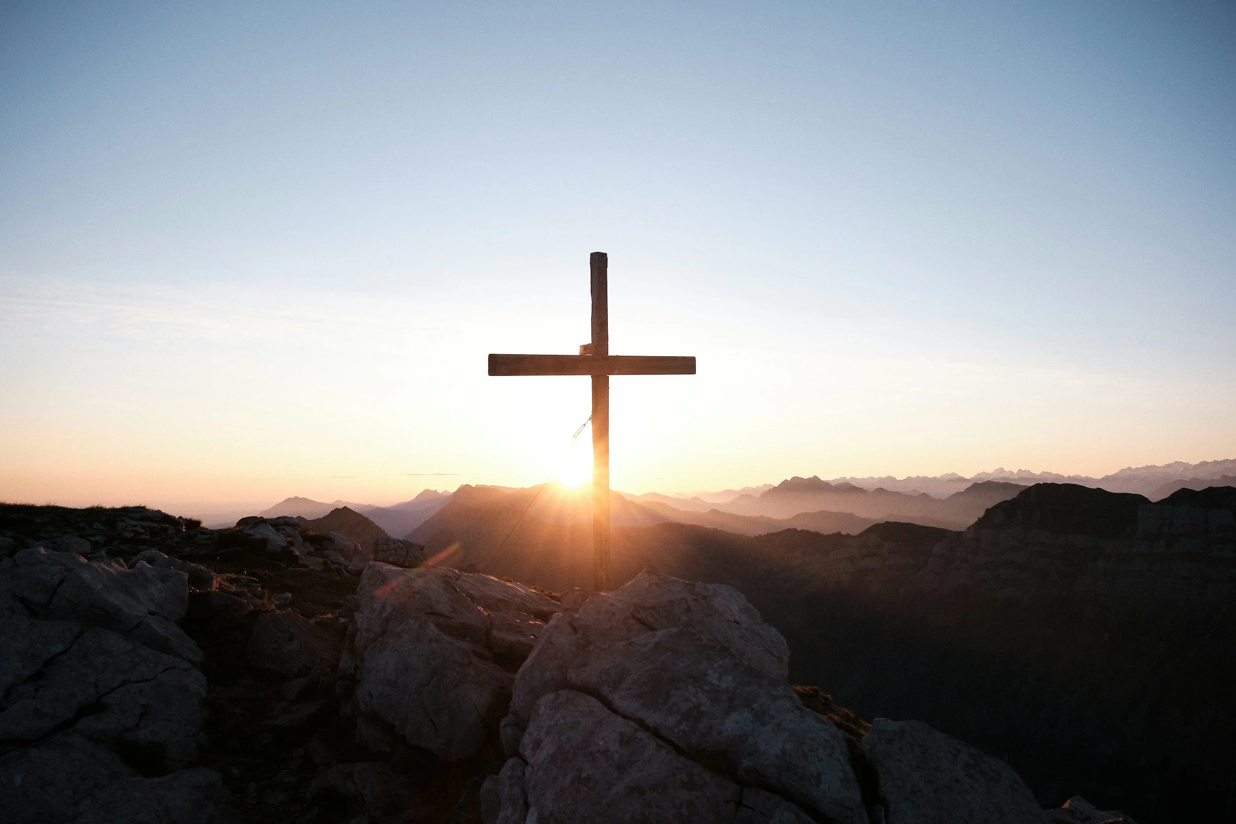 A wooden cross stands on a rocky mountaintop against a sunrise or sunset sky, with distant mountain ranges in the background.