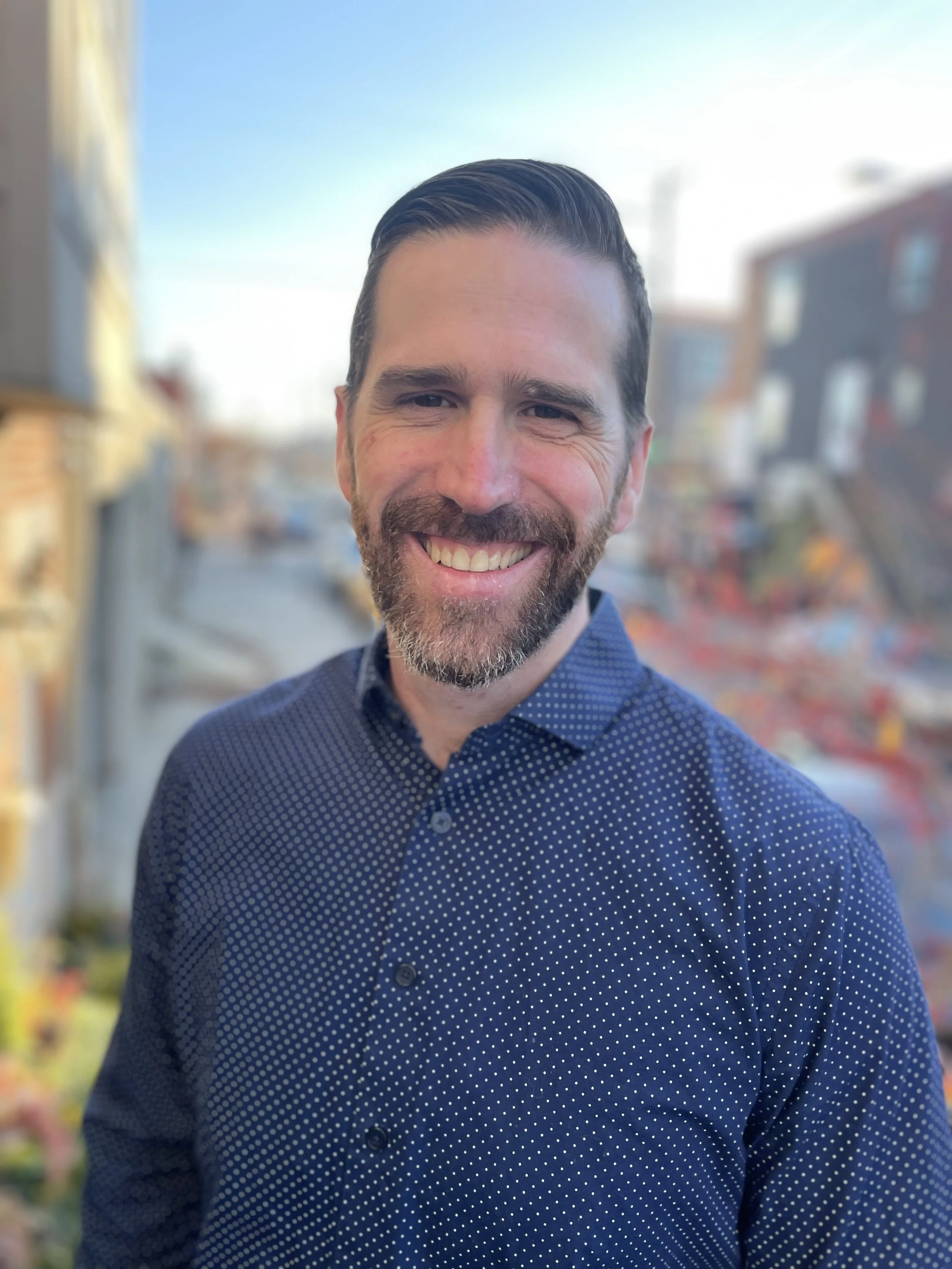 A man with dark hair and a beard smiling, wearing a navy blue shirt with white polka dots, standing outdoors with blurred buildings and trees in the background.