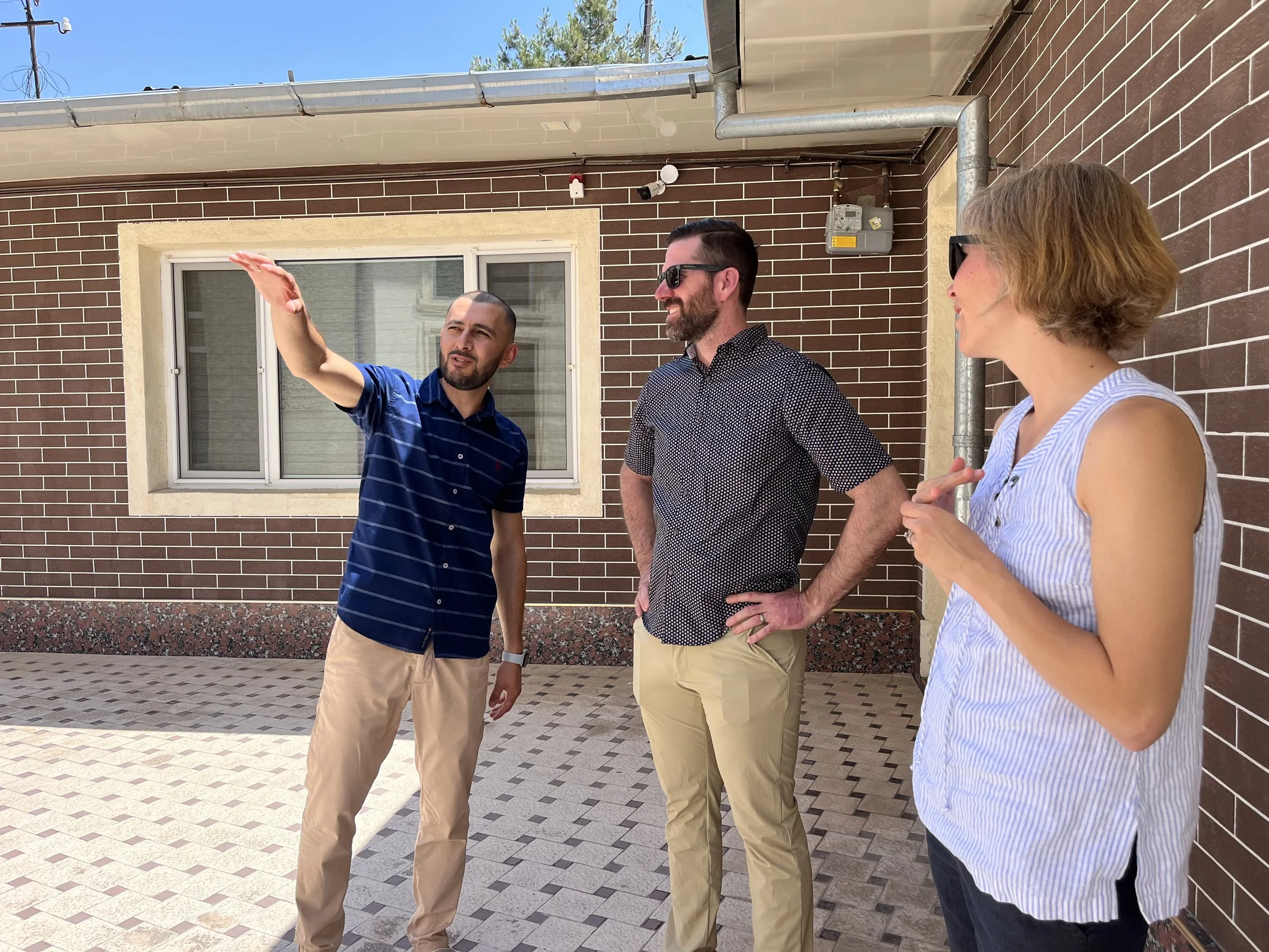 Three people standing and talking outside a brick building, with a man in a blue striped shirt gesturing with his hand, and two others, one a bearded man wearing sunglasses and a patterned shirt, and a woman with short hair and glasses, listening attentively.