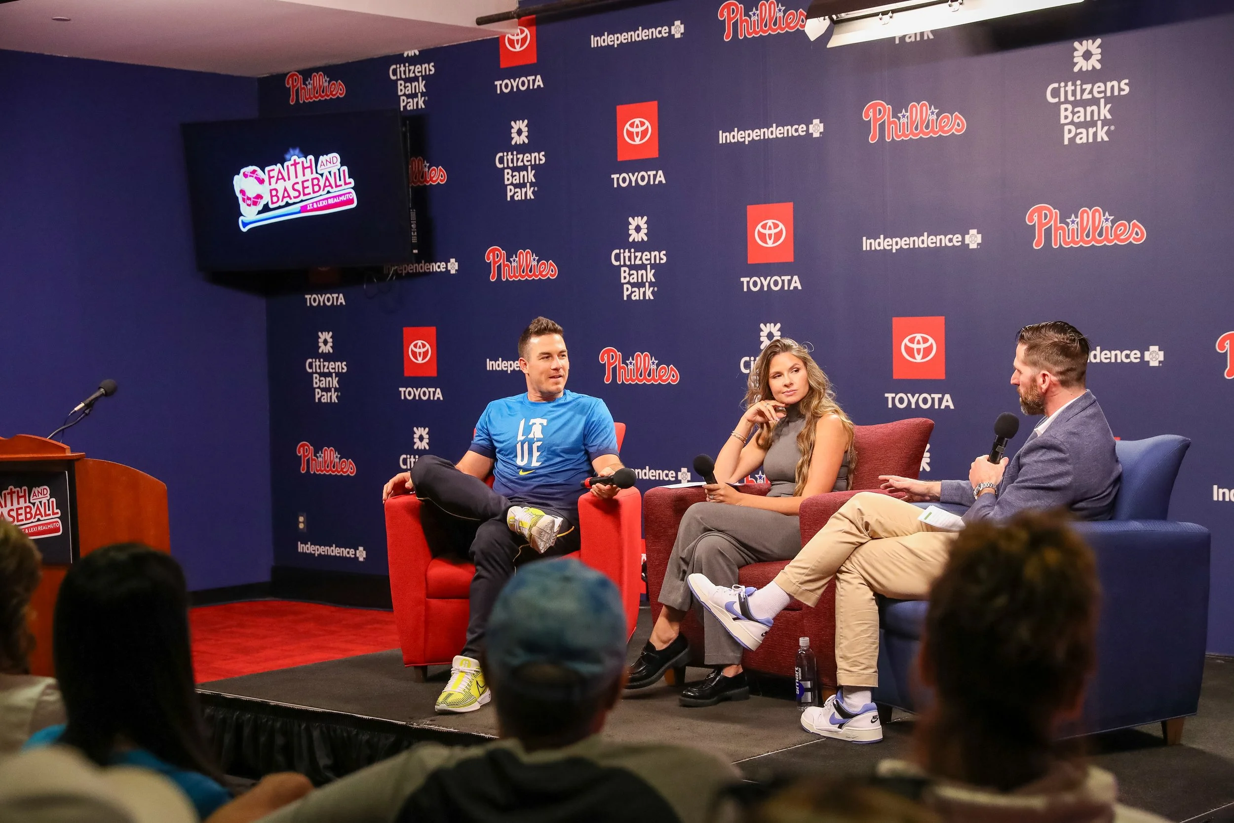 A panel discussion taking place at Citizens Bank Park with three individuals seated on stage in front of a blue backdrop featuring various sponsor logos. The backdrop has logos for the Phillies, Toyota, Citizens Bank Park, and Independence. There is a large screen displaying the Faith and Baseball logo, and the audience is visible in the foreground.