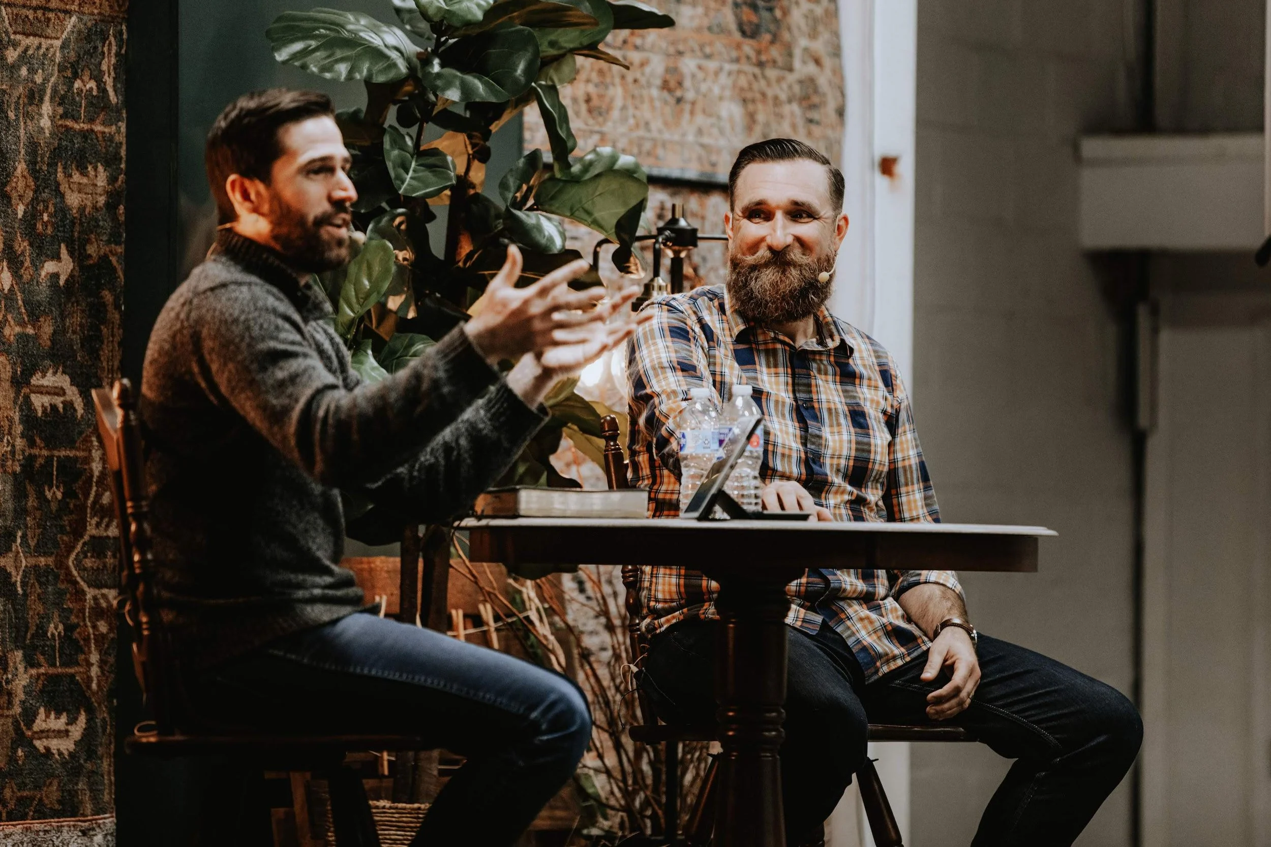Two men engaged in a conversation, sitting at a table in an indoor setting with a large plant and brick wall behind them. One man with a beard and plaid shirt is smiling, the other with dark hair and gray sweater is gesturing.