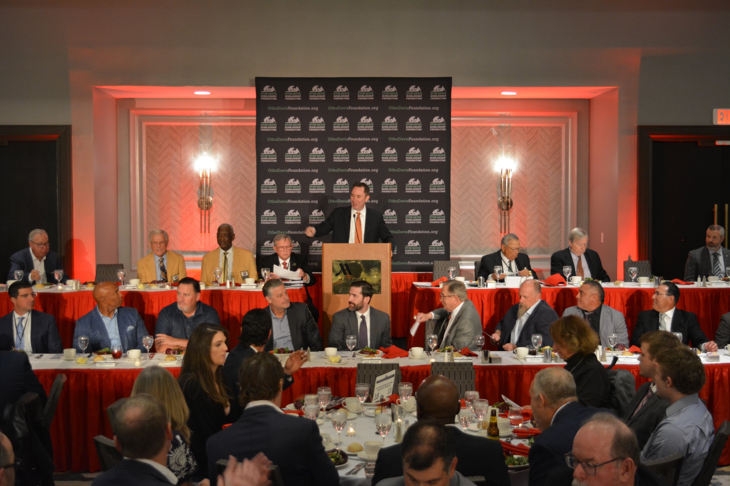 A man in a black suit and orange tie is standing at a podium speaking to an audience at a formal event, with a black backdrop displaying the Otho Davis Foundation logo behind him. Seated at a long table behind him are several men in suits, some taking notes. The room is decorated with orange lighting and wall sconces.