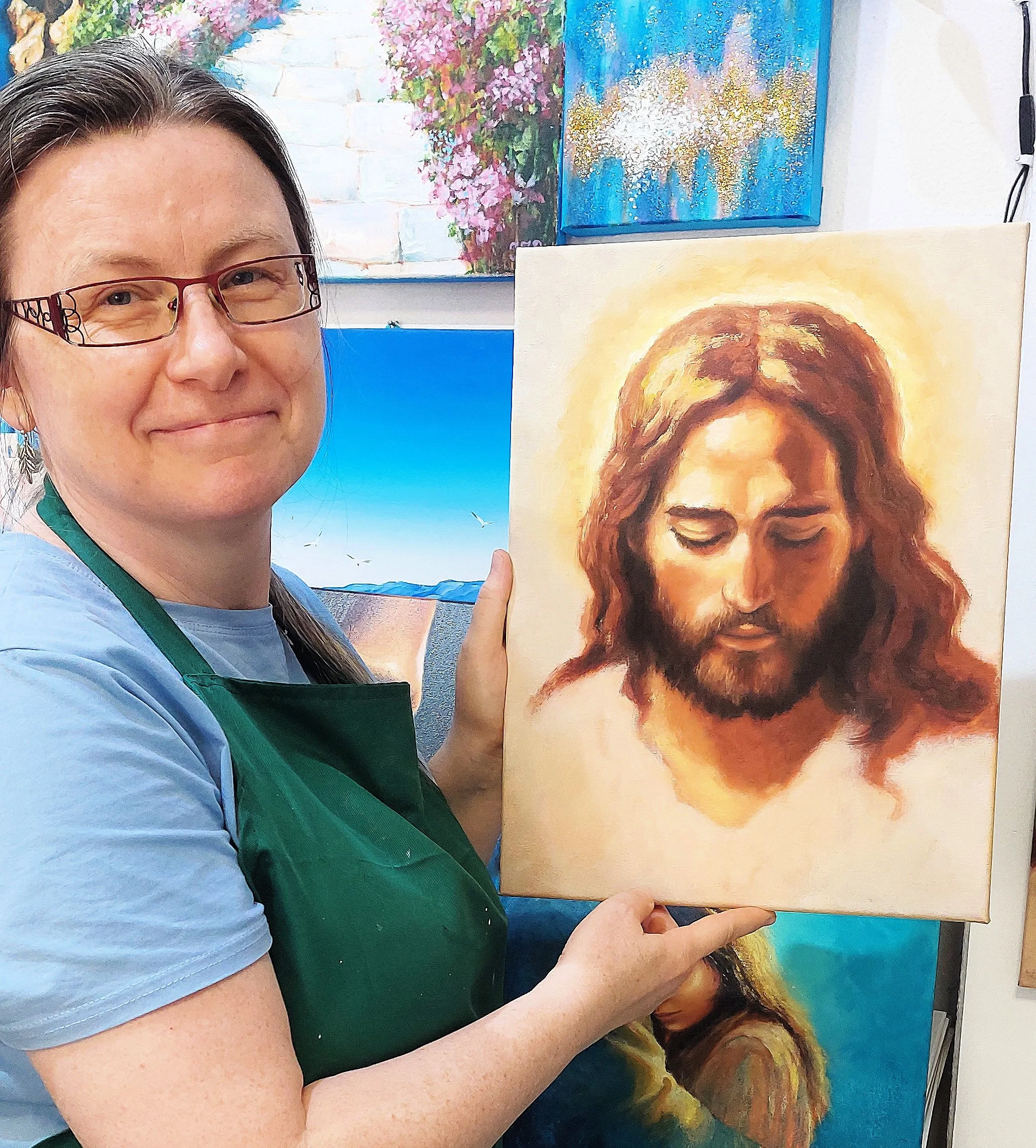 A woman wearing glasses and a green apron holding an oil painting of Jesus Christ with closed eyes and brown hair. There are colorful landscape paintings hanging on the wall in the background.