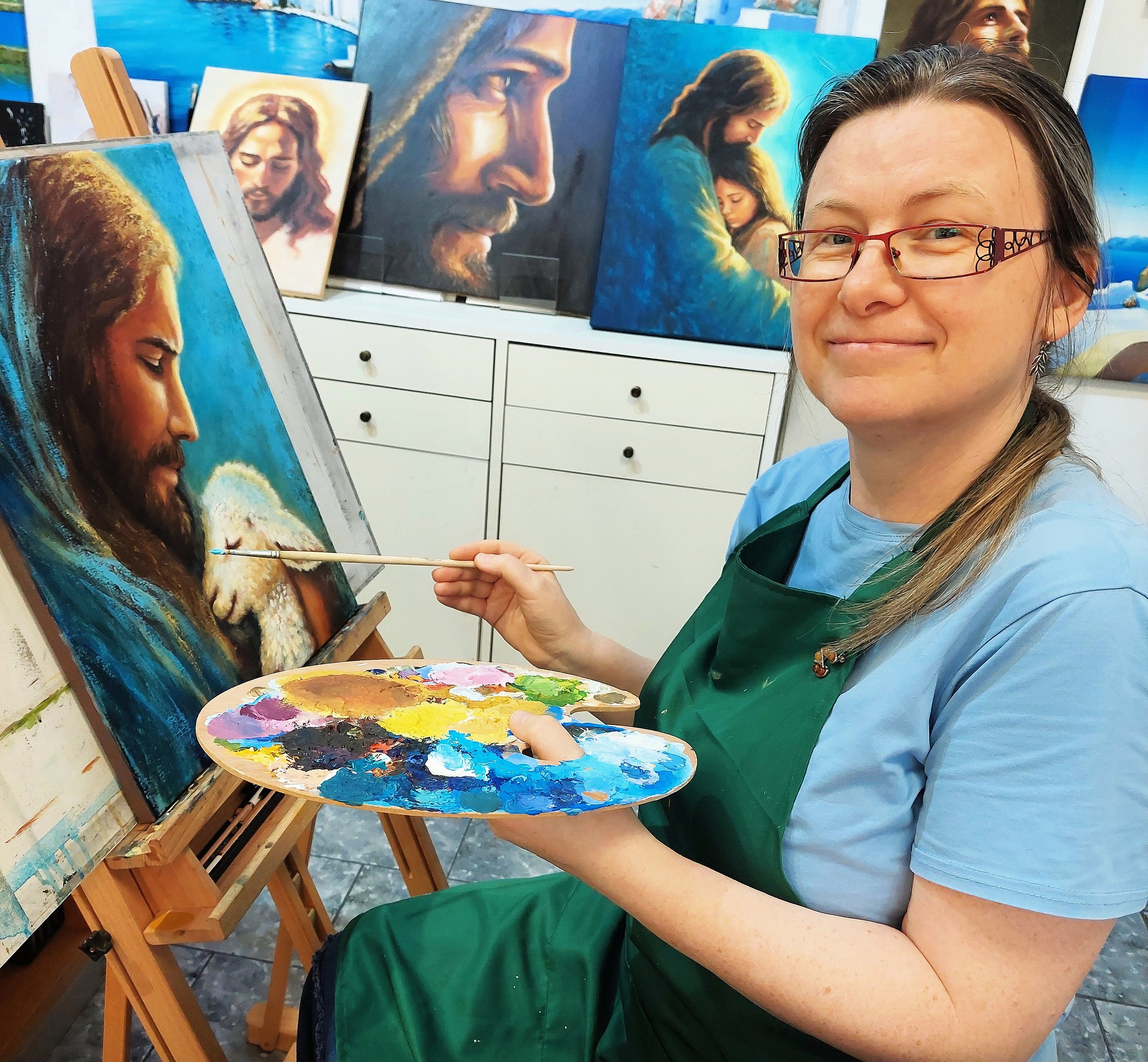 A young woman with glasses and a ponytail smiling at the camera while holding a palette of mixed paints. She is wearing a blue shirt and green apron, working on a painting of Jesus with long hair and a beard. Several other paintings of the same subject are displayed on easels behind her.