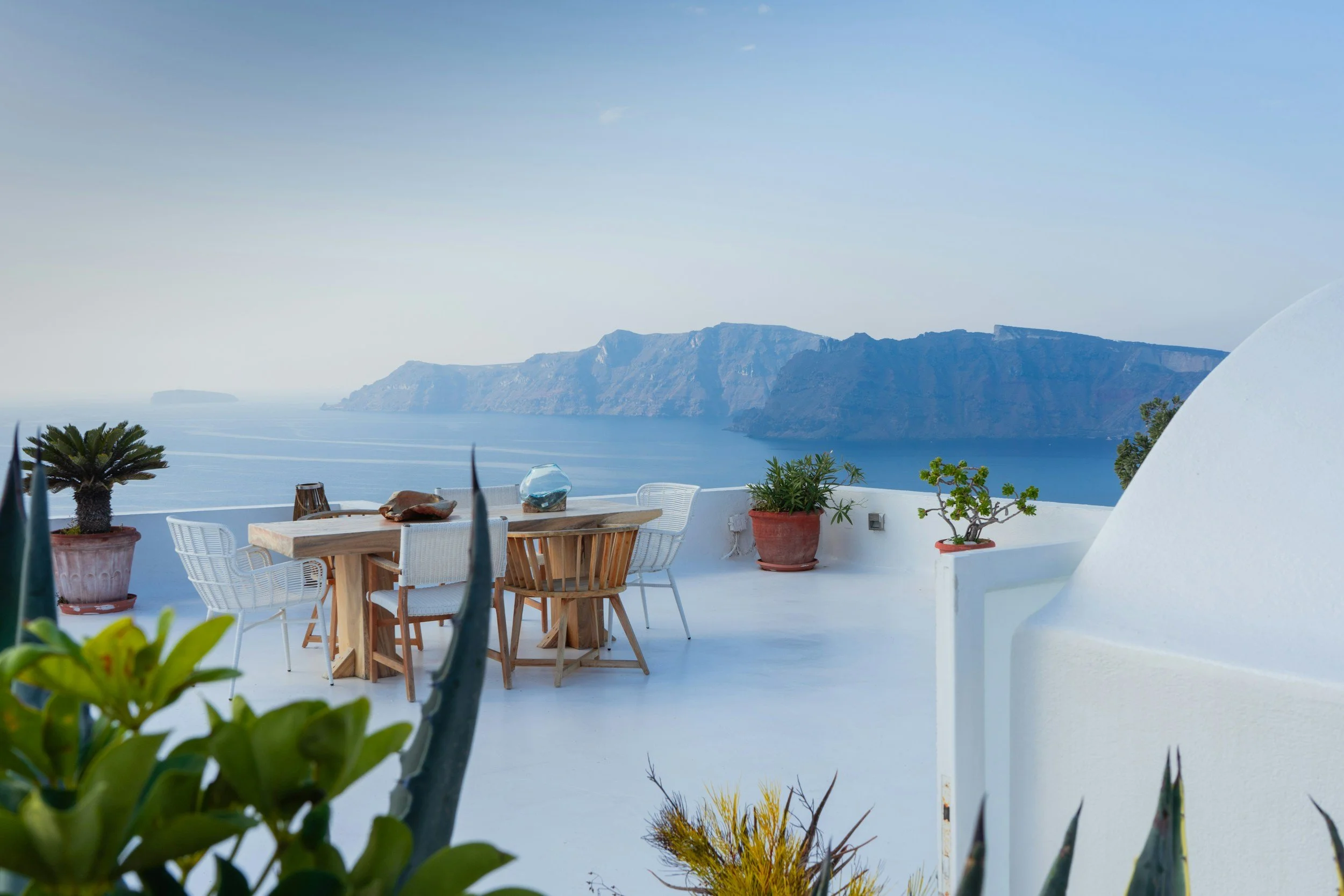 A scenic view of a terrace with potted plants and a wooden table surrounded by white chairs, overlooking a calm blue sea and distant cliffs under a clear sky.