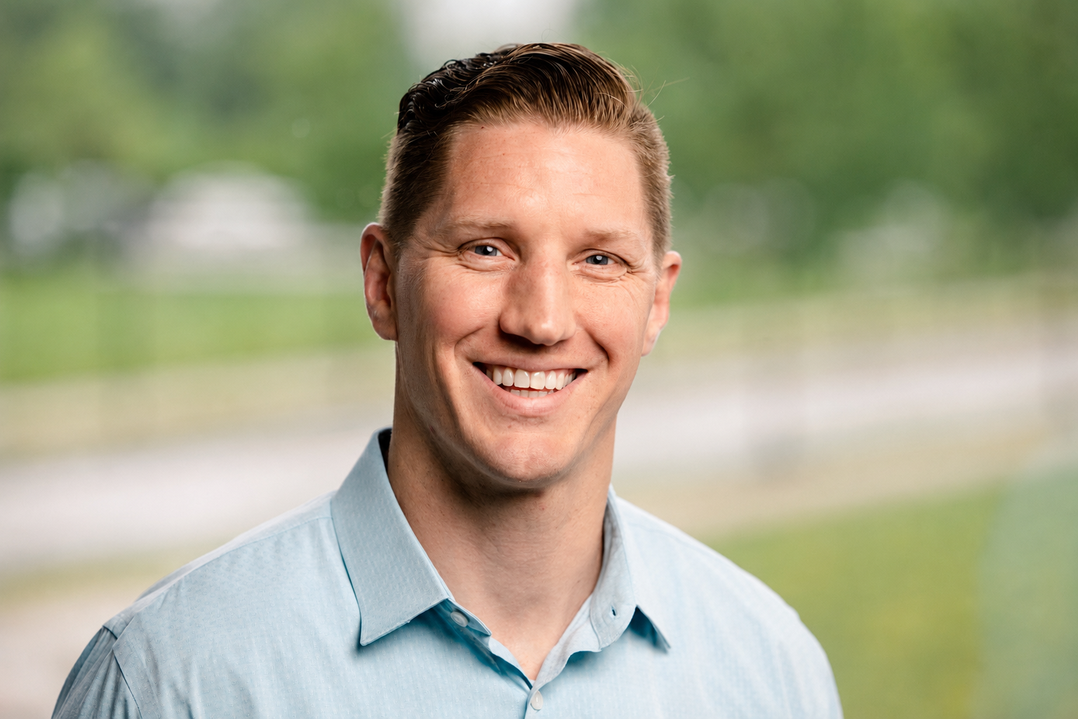 A smiling man with short, neatly styled brown hair wearing a light blue collared shirt, standing outdoors with a blurred green landscape in the background.
