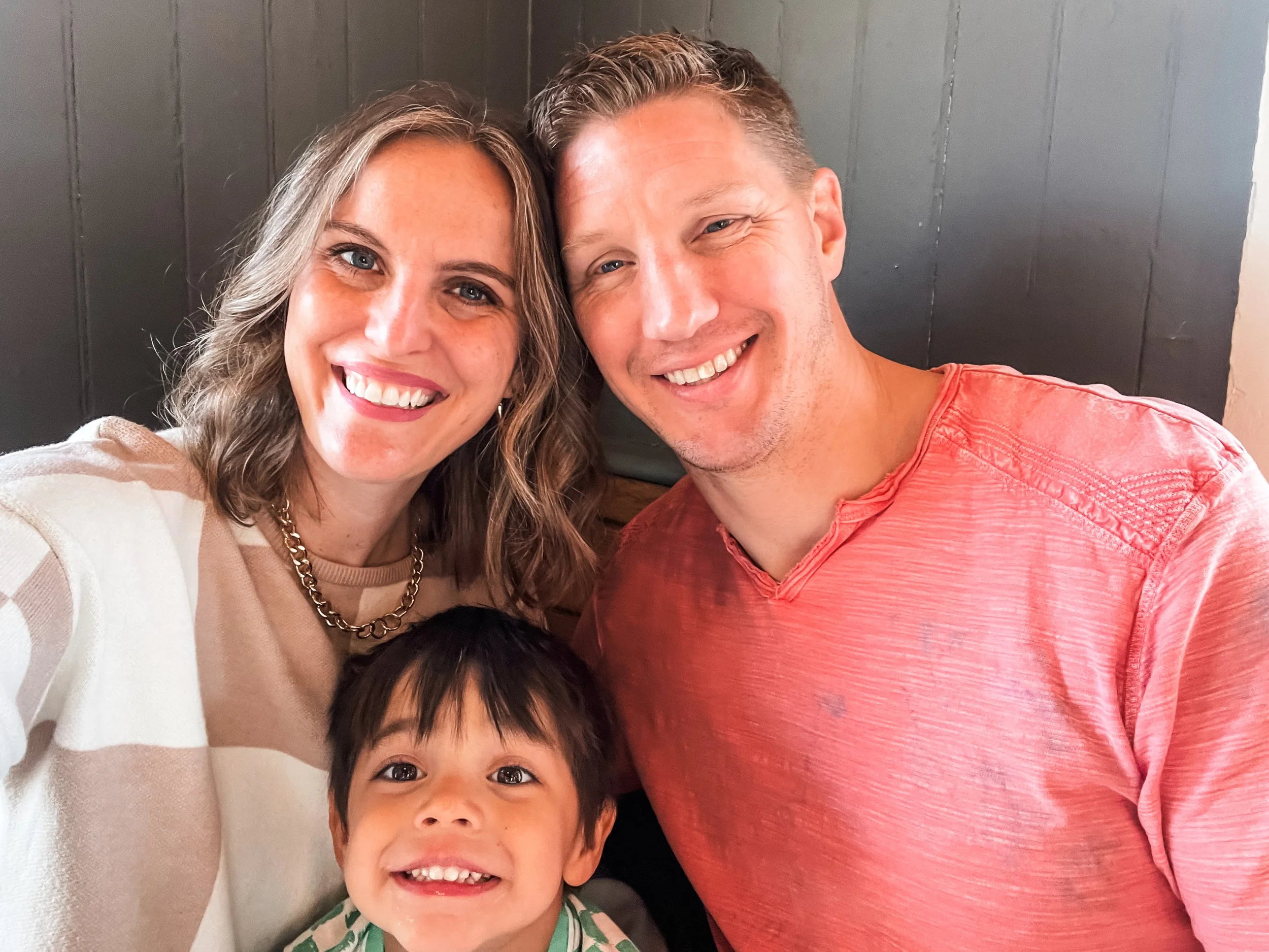 A smiling woman with long, light brown hair and a beige striped top, a young boy with dark hair smiling widely, and a man with short light brown hair wearing a red shirt, all posing together indoors against a dark wall with vertical panels.