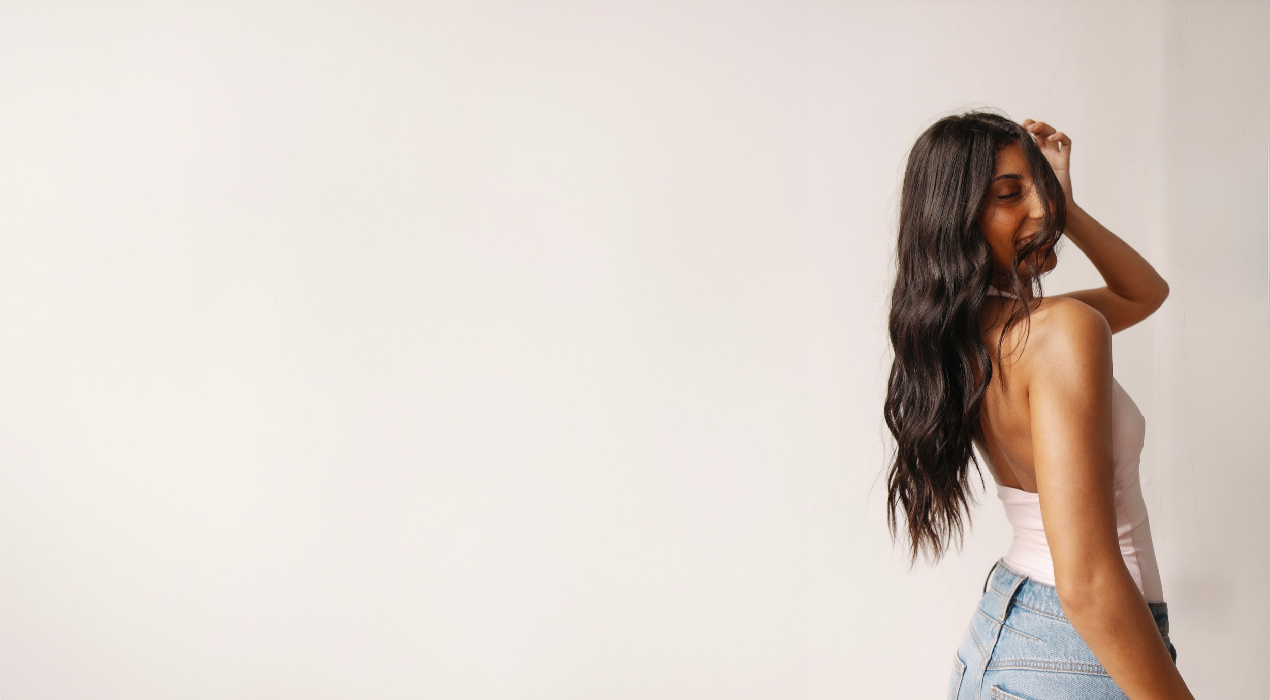 Young woman with long, wavy dark hair smiling and posing in front of a plain light-colored wall, wearing a light pink tank top and jeans.