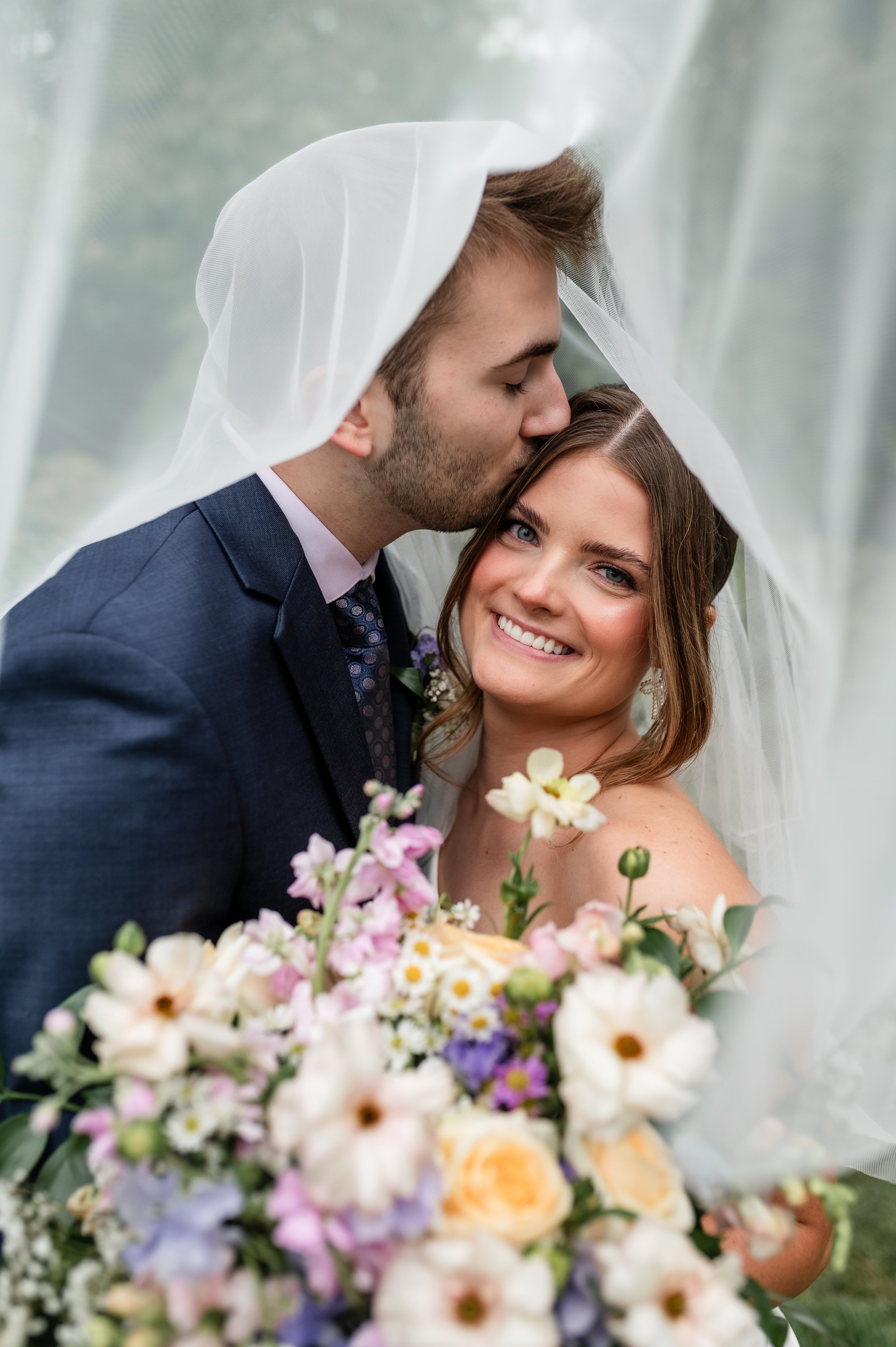 Groom kisses bride on the forehead under a veil; bride smiles with makeup done by a wedding makeup artist holding a bouquet of pastel flowers.