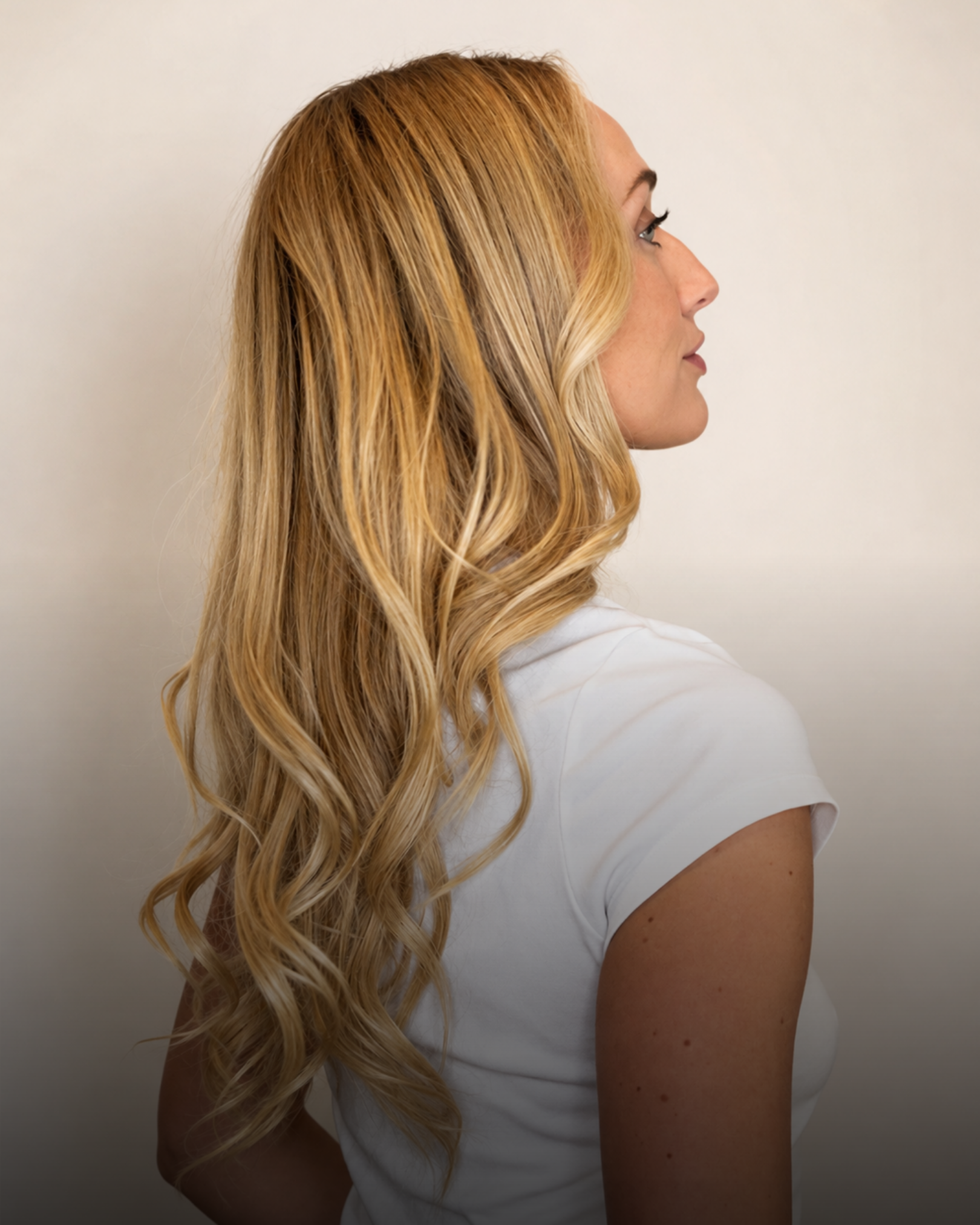 Woman with a blowout for long, wavy blonde extensions wearing a white shirt, shown in profile against a neutral background.