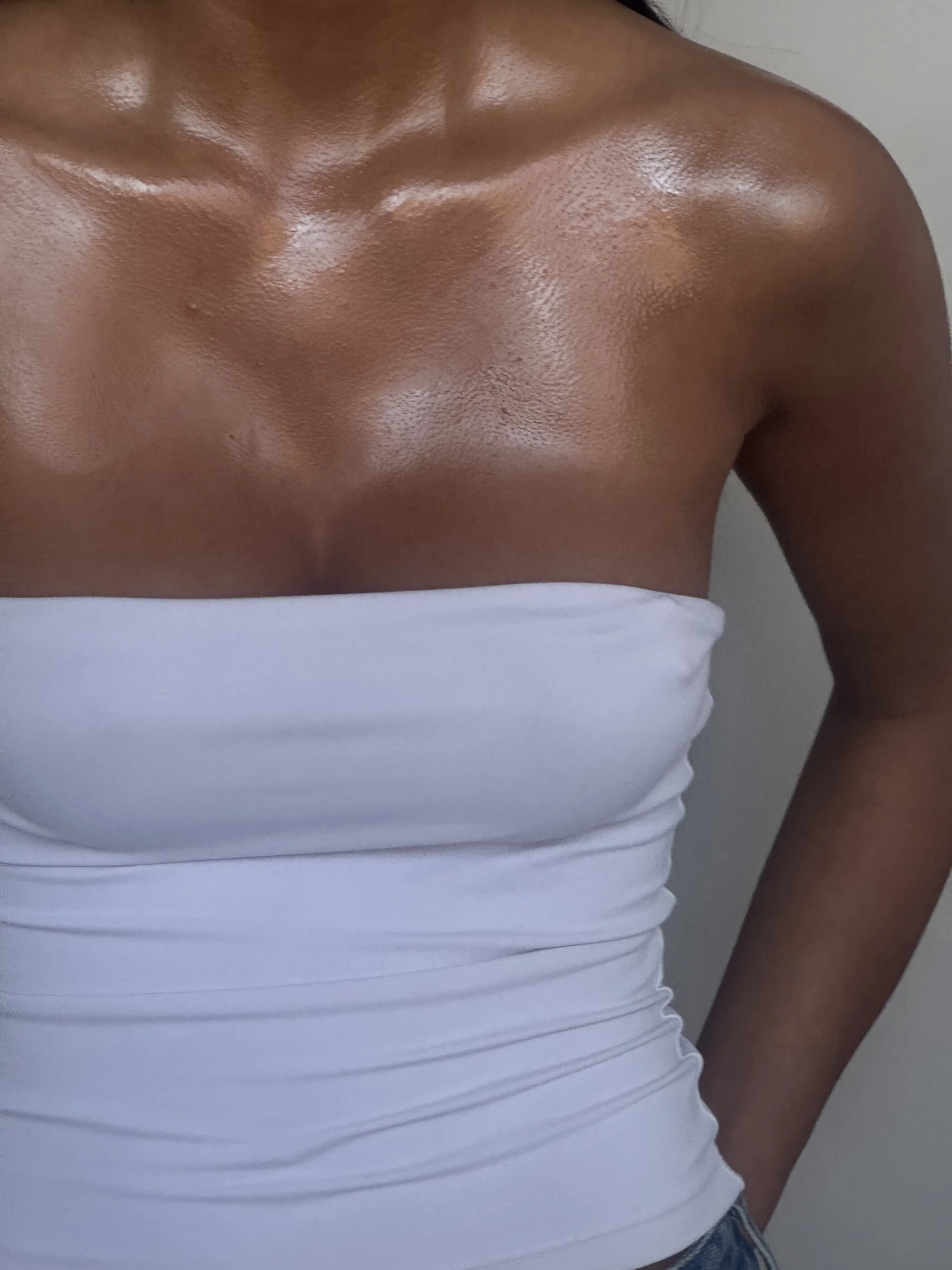 Person wearing a white strapless top with shiny, custom airbrush tanned skin visible on their chest and shoulders.