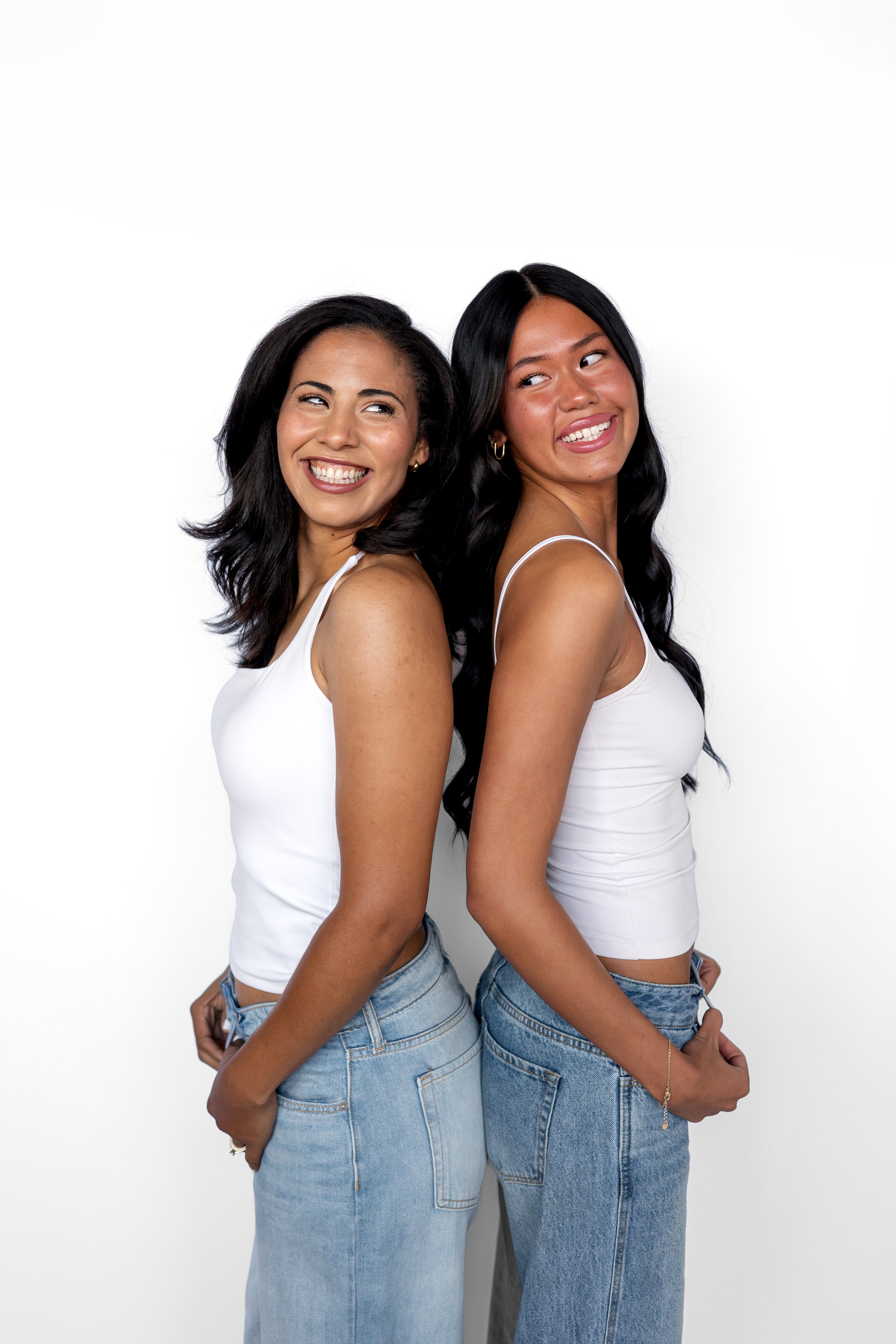 Two women stand back-to-back, smiling in a beauty salon in Niles, Ohio, dressed in white tops and blue jeans against a plain white background.
