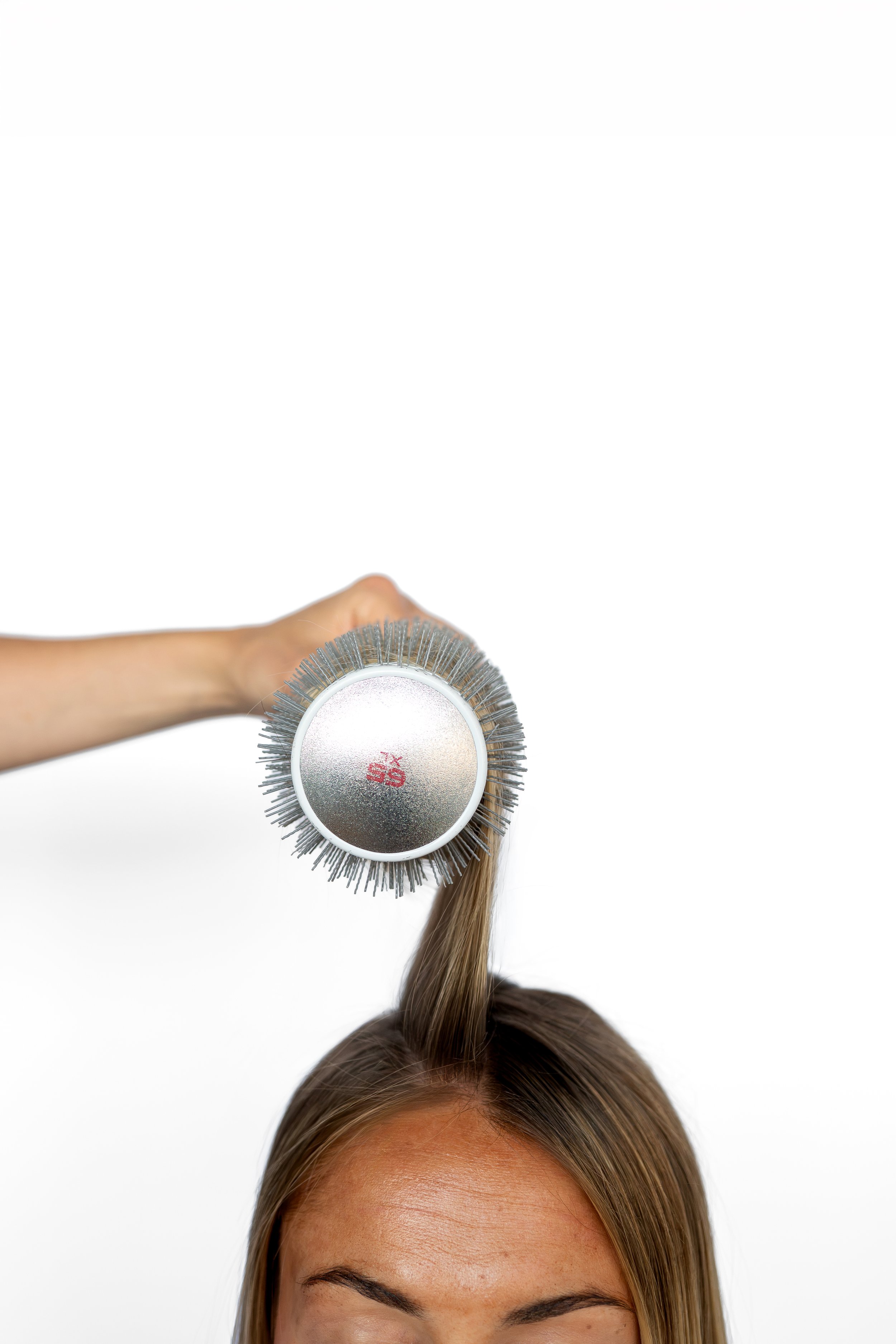Person holding a round hairbrush and lifting a section of straight brown hair from the top of their head performing a blowout at a blow dry bar.