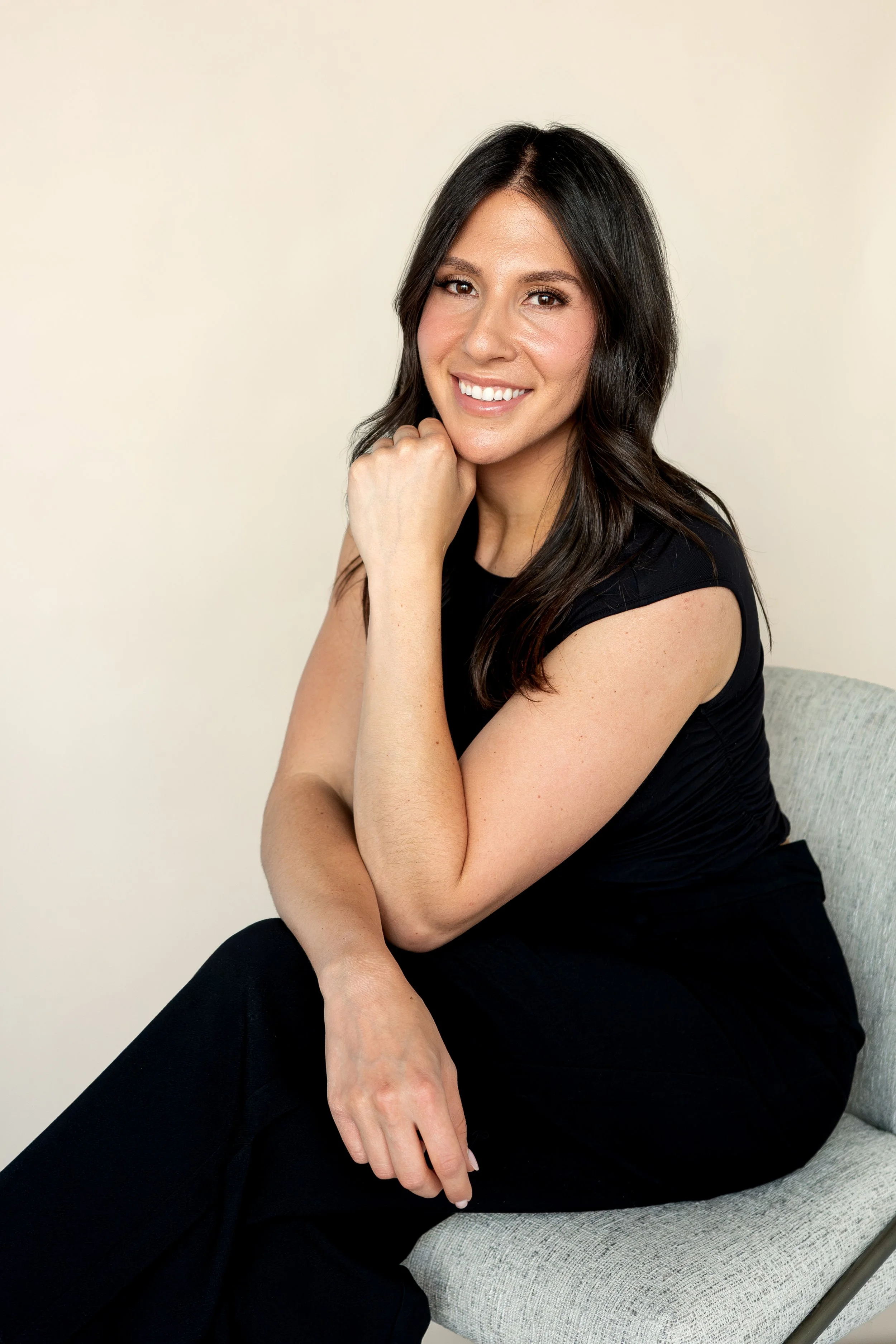 Karsyn Berk, the founder of hello blow, with long dark hair, wearing a black outfit, sits on a light chair and smiles at the camera against a plain background.