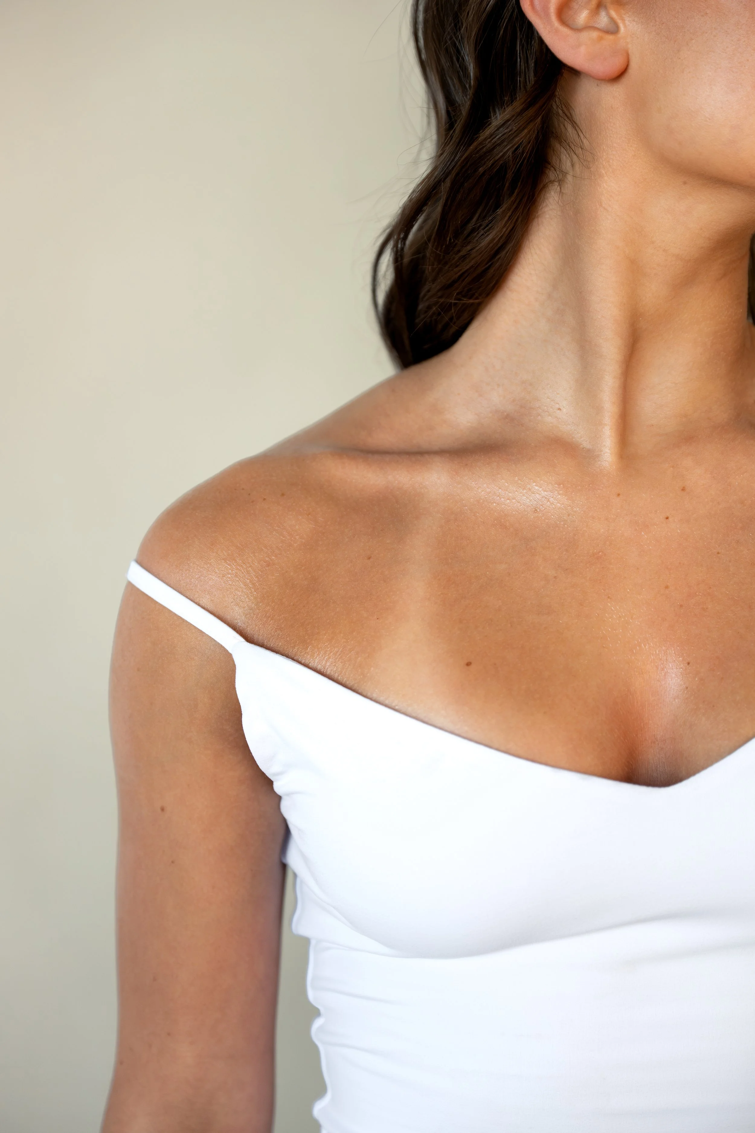 Woman wearing a white spaghetti strap top, showing her custom spray tan against a plain background.