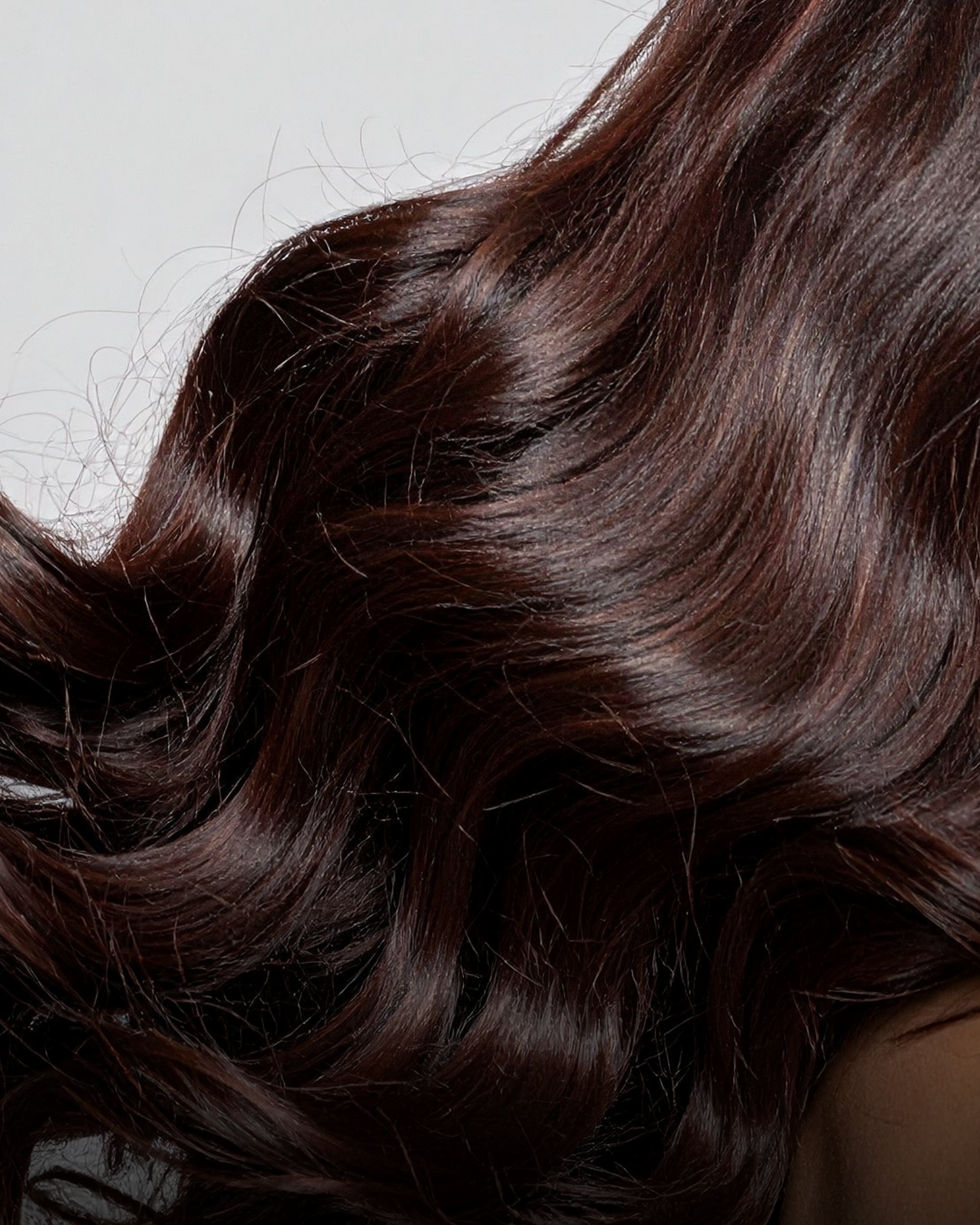 Close-up of long, wavy, dark brown hair with a glossy texture from a blow dry salon, against a plain light background.