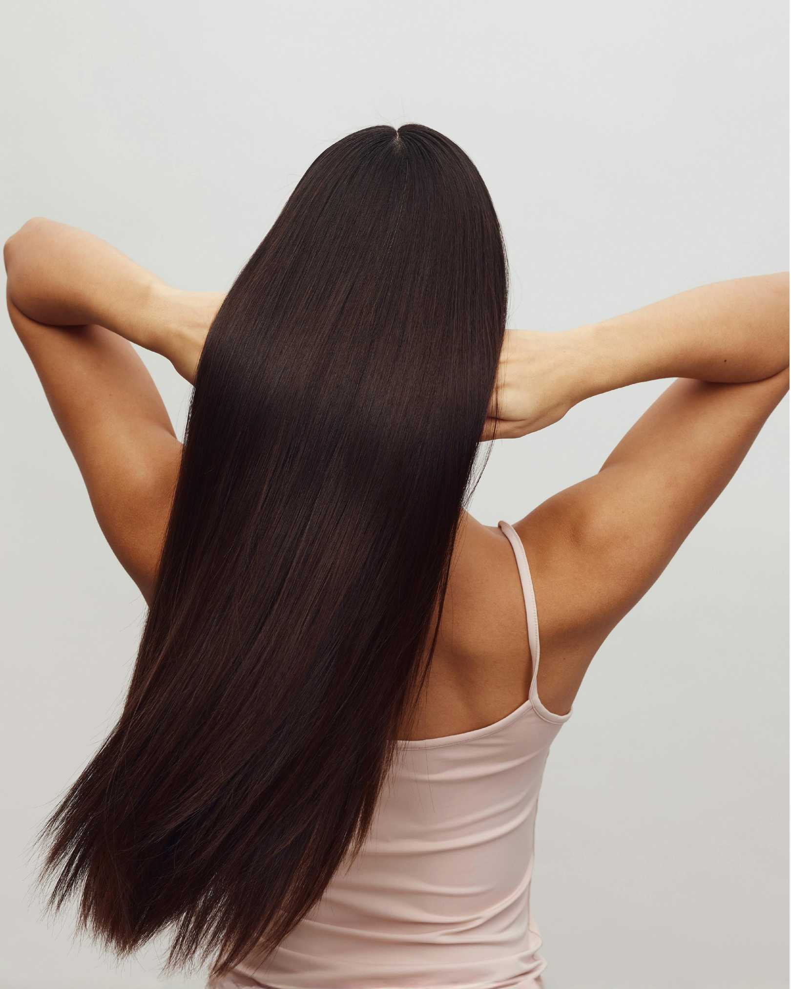 Person with long, straight, dark brown extensions shown from the back, hands behind neck, wearing a light tank top.