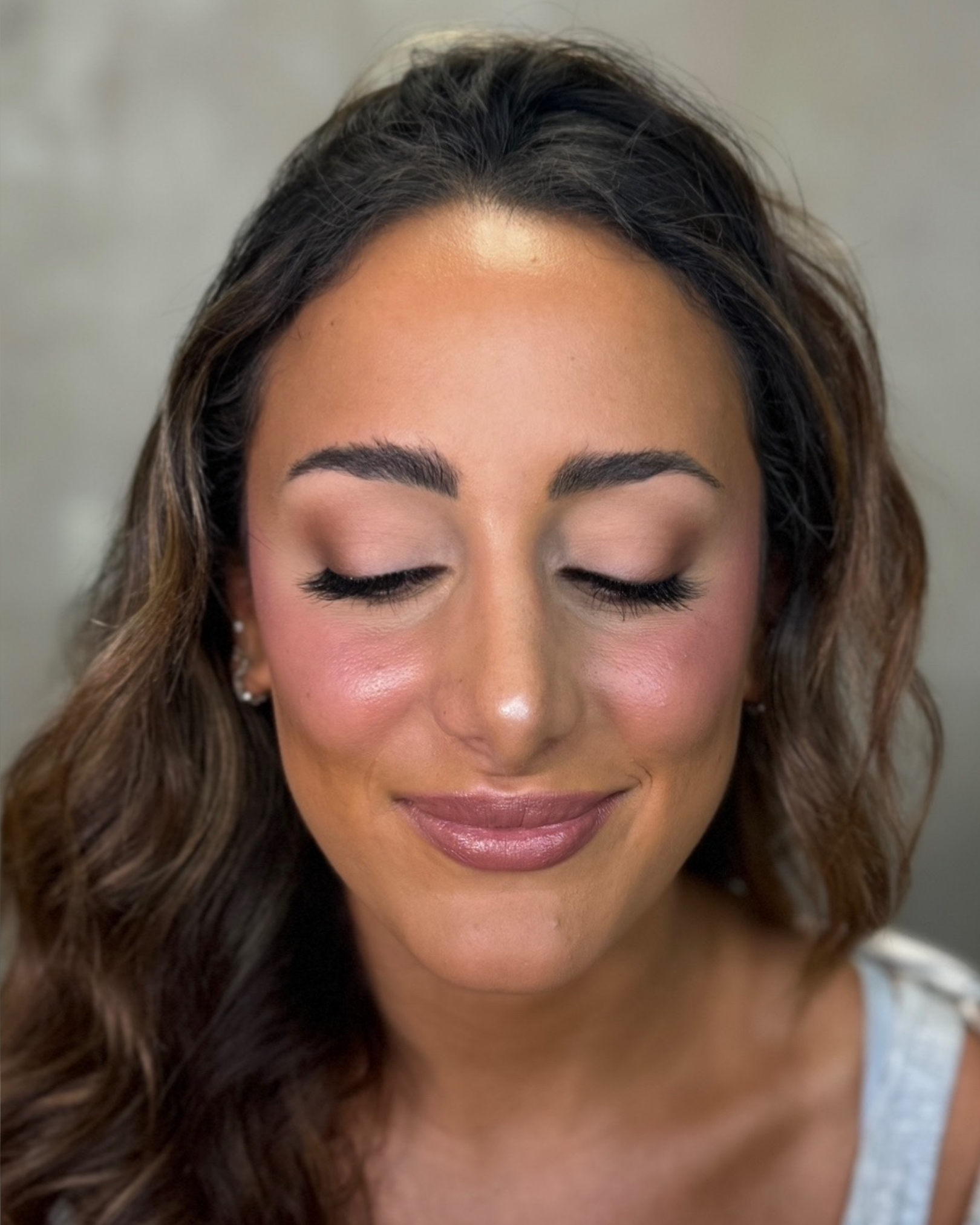 Woman with long brown hair and soft eye glam makeup smiles with her eyes closed against a neutral background.