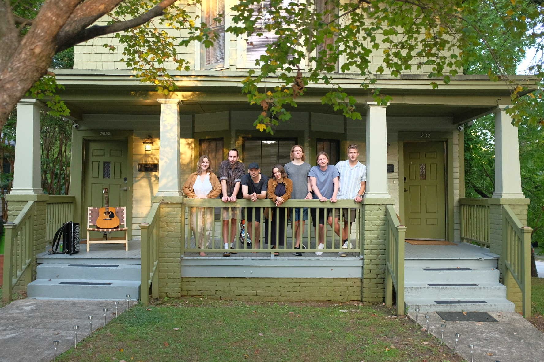 Group of seven people standing on the porch of a green house with a guitar and a black bag on a chair, surrounded by trees.