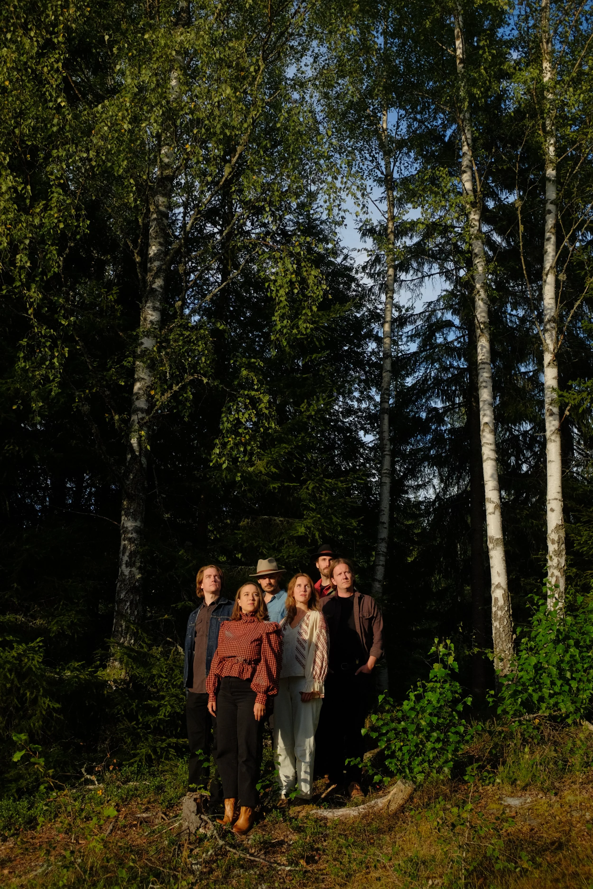 The band consisting of six people, four women and two men, are standing in a forest with tall trees, some with white bark, and dense greenery. They are looking in different directions, and the sunlight is filtering through the trees.