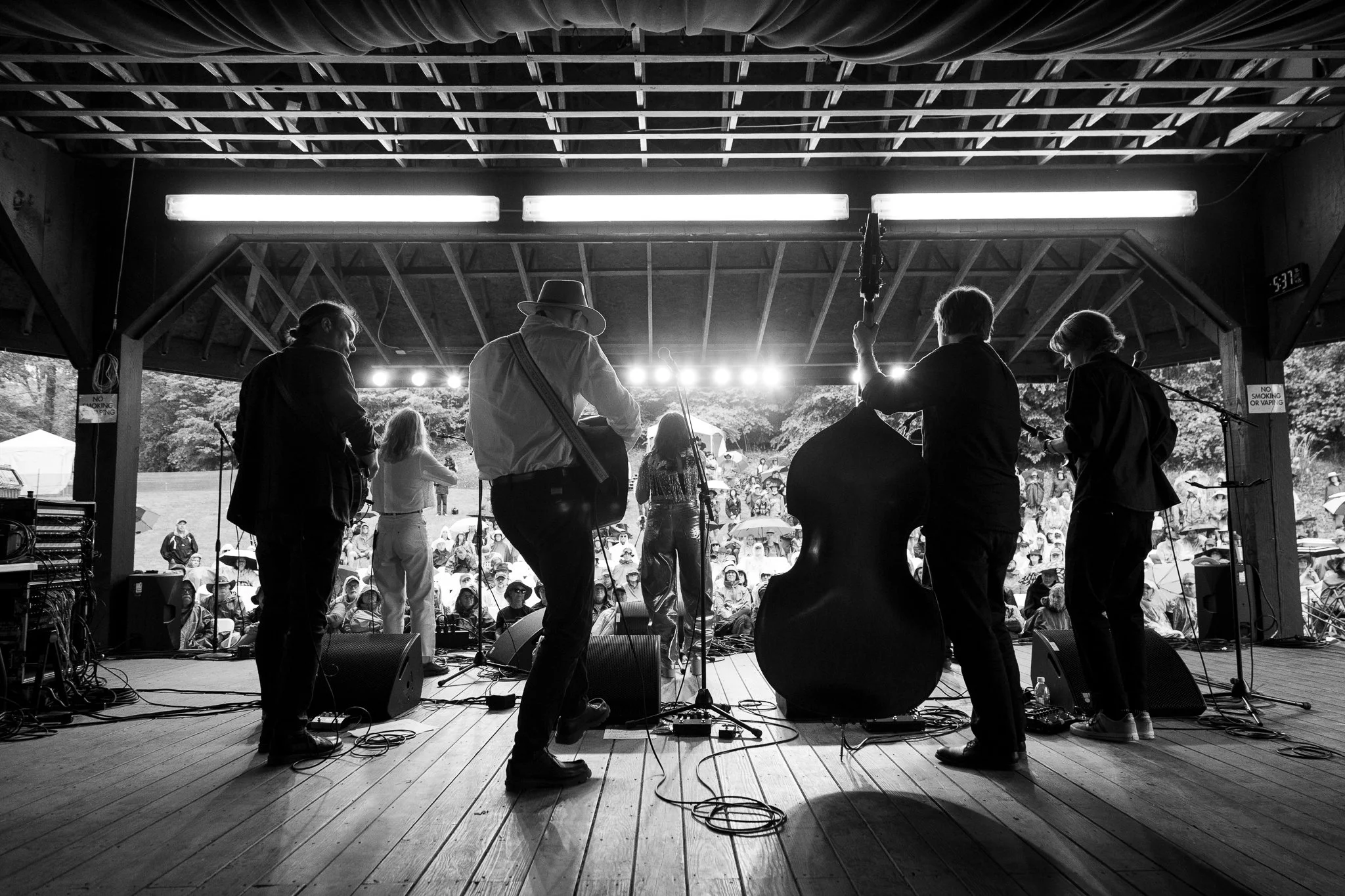 Black and white photo of a musical performance on an outdoor stage, with the audience in view. The band is seen from behind.