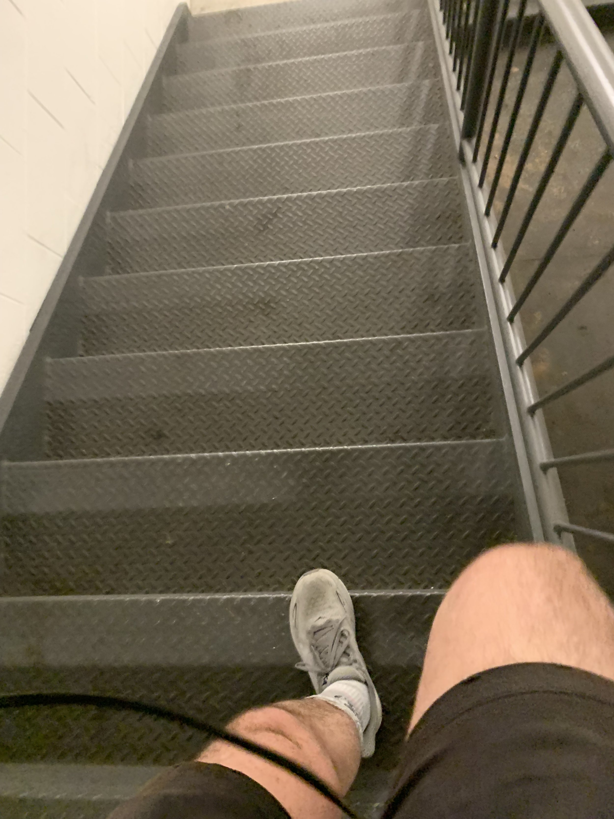 A person's legs and feet wearing white sneakers, standing on metal stairs with diamond plate pattern, with a black metal railing on the right side.