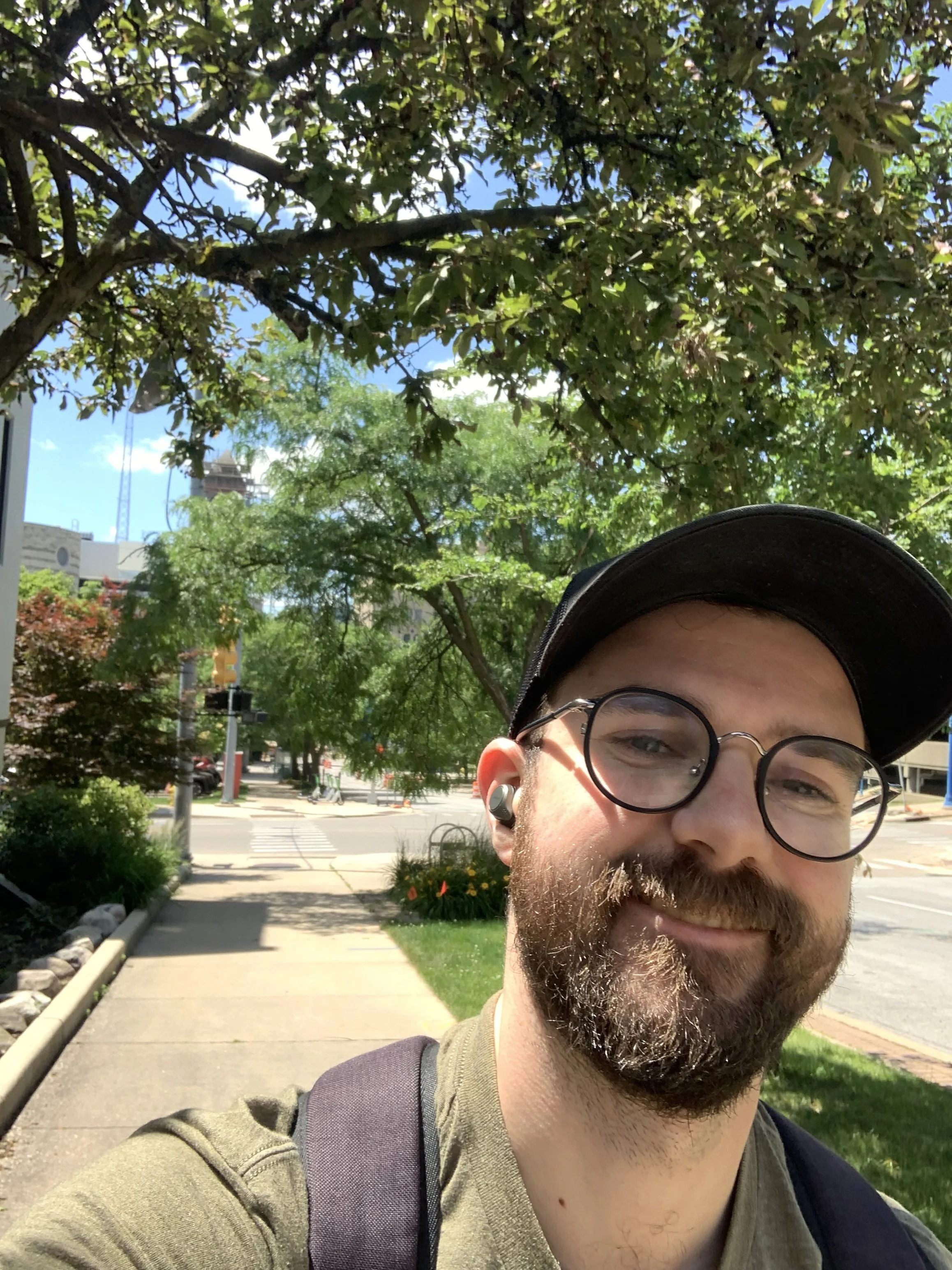 A man with glasses and a beard smiling outdoors in a sunny park, wearing a hat and backpack, with trees and a city street in the background.