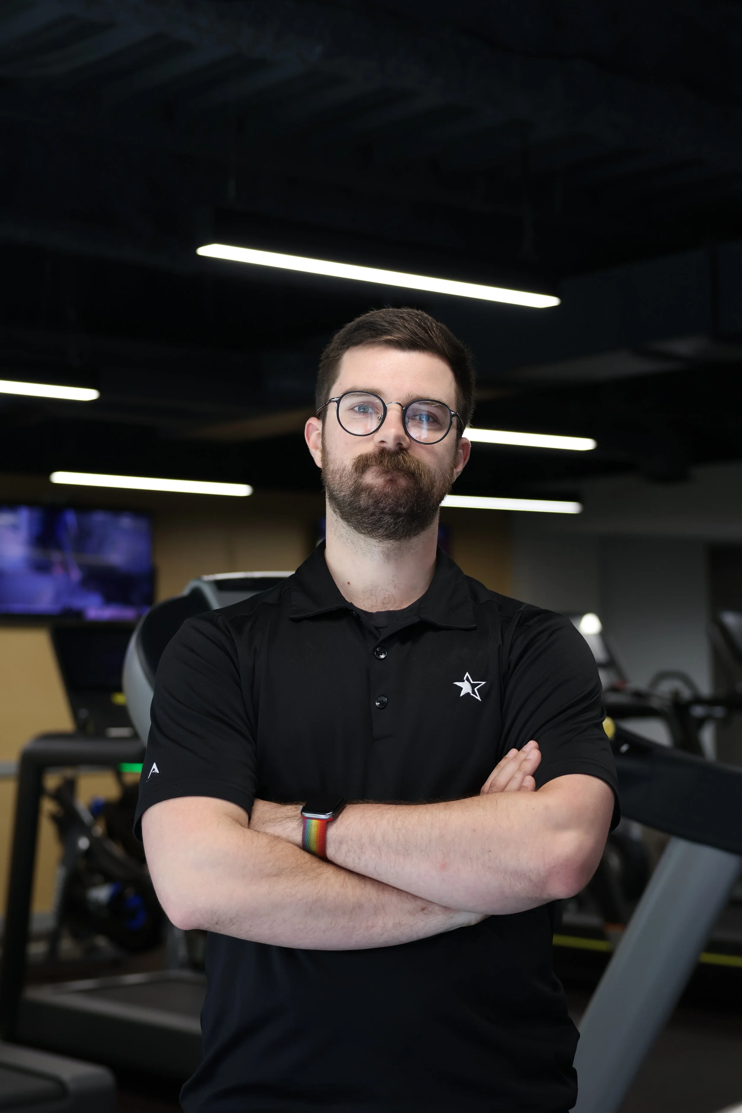 A fit man with glasses, a beard, and mustache standing with arms crossed in a gym, wearing a black polo shirt and a rainbow wristband, in front of exercise equipment, with dark ceiling and fluorescent lights above.