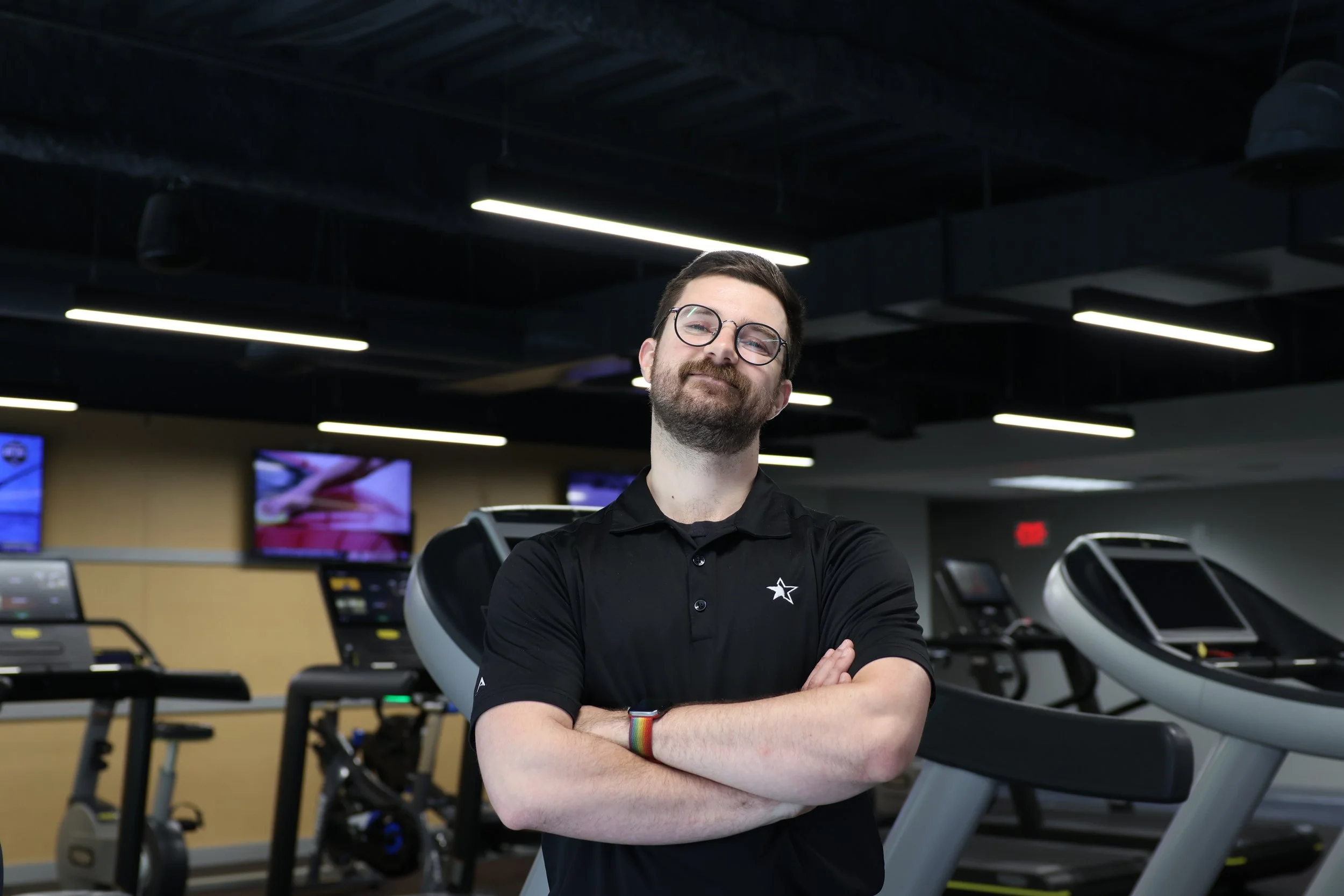 A man with glasses and a beard standing with arms crossed in a gym with treadmills.