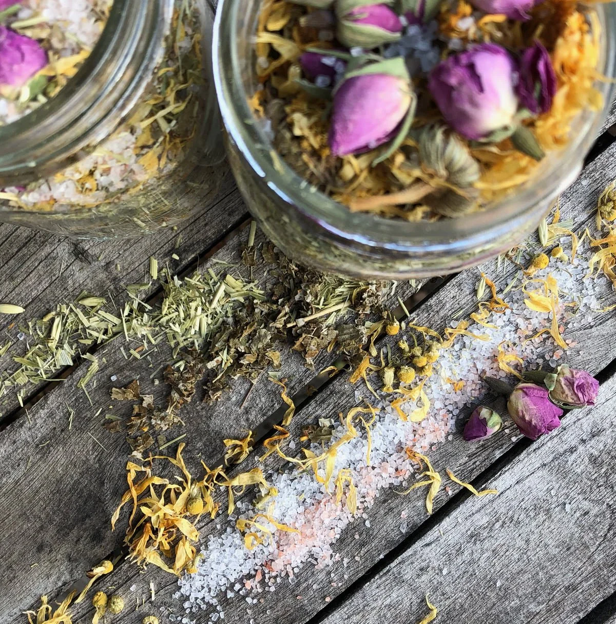 Two glass jars filled with dried herbs and flower petals, with some herbs and flower petals spilled on a rustic wooden surface underneath. The jars contain dried rose buds and other botanical ingredients.