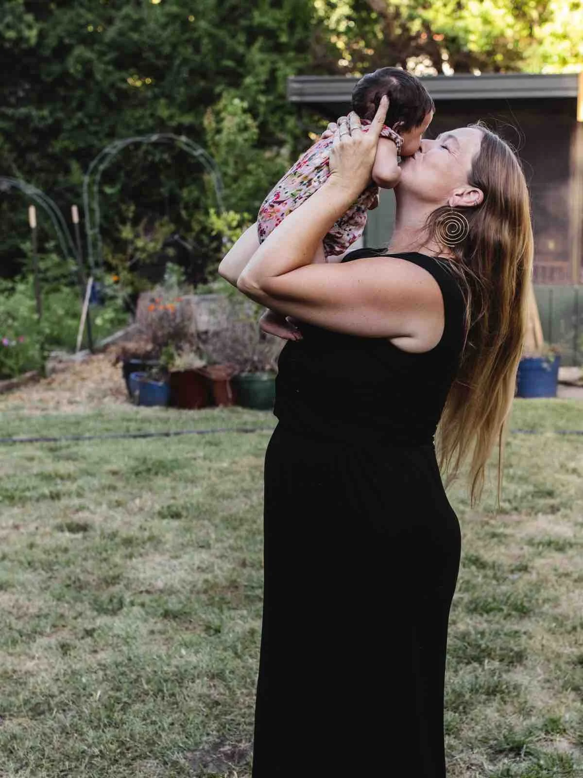 A woman in a black dress holding a young girl and kissing her outdoors in a backyard with trees and garden beds.