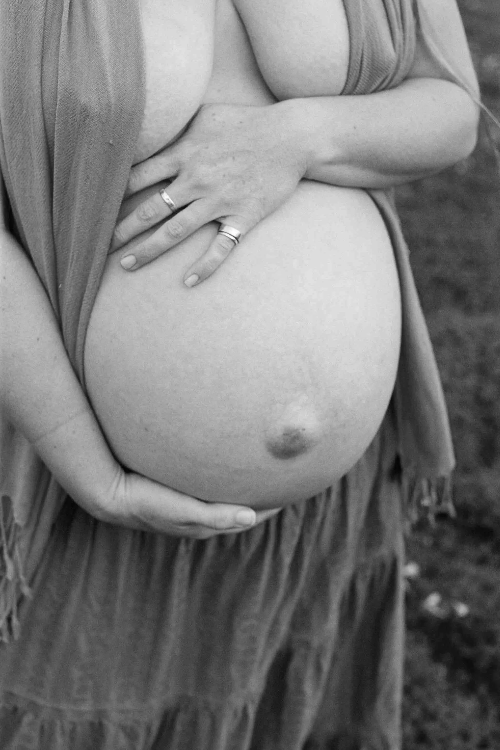 A close-up black and white photo of a pregnant woman cradling her belly with her hands, wearing rings on her fingers and a sleeveless top, with a background that appears to be outdoors.