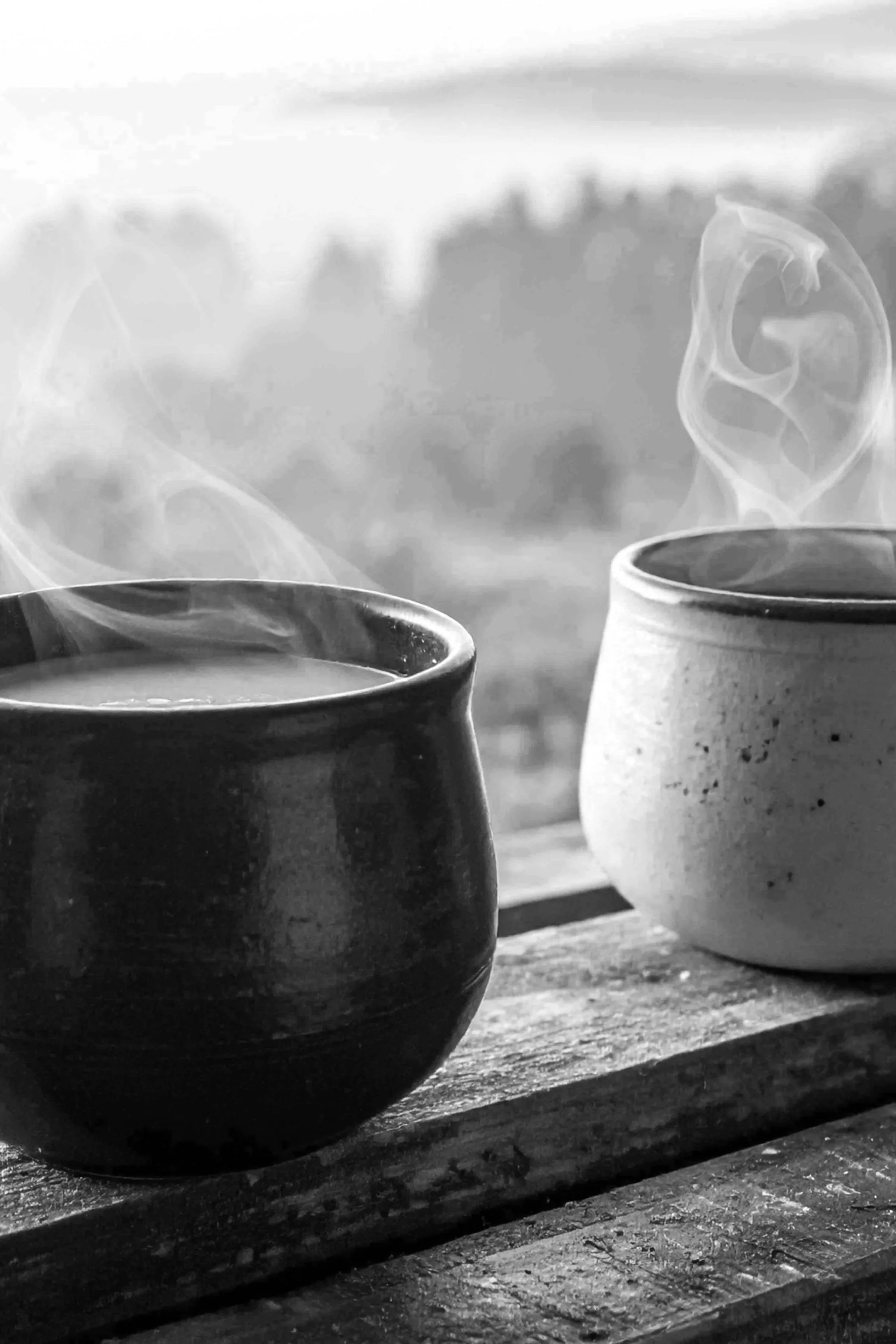 Two steaming cups on a wooden surface with a blurred outdoor background in black and white.