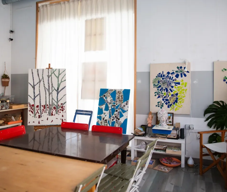 An art studio with three mosaic paintings of trees and foliage displayed on easels and the wall, with a table and chairs in the foreground and various art supplies and decorations around the room.