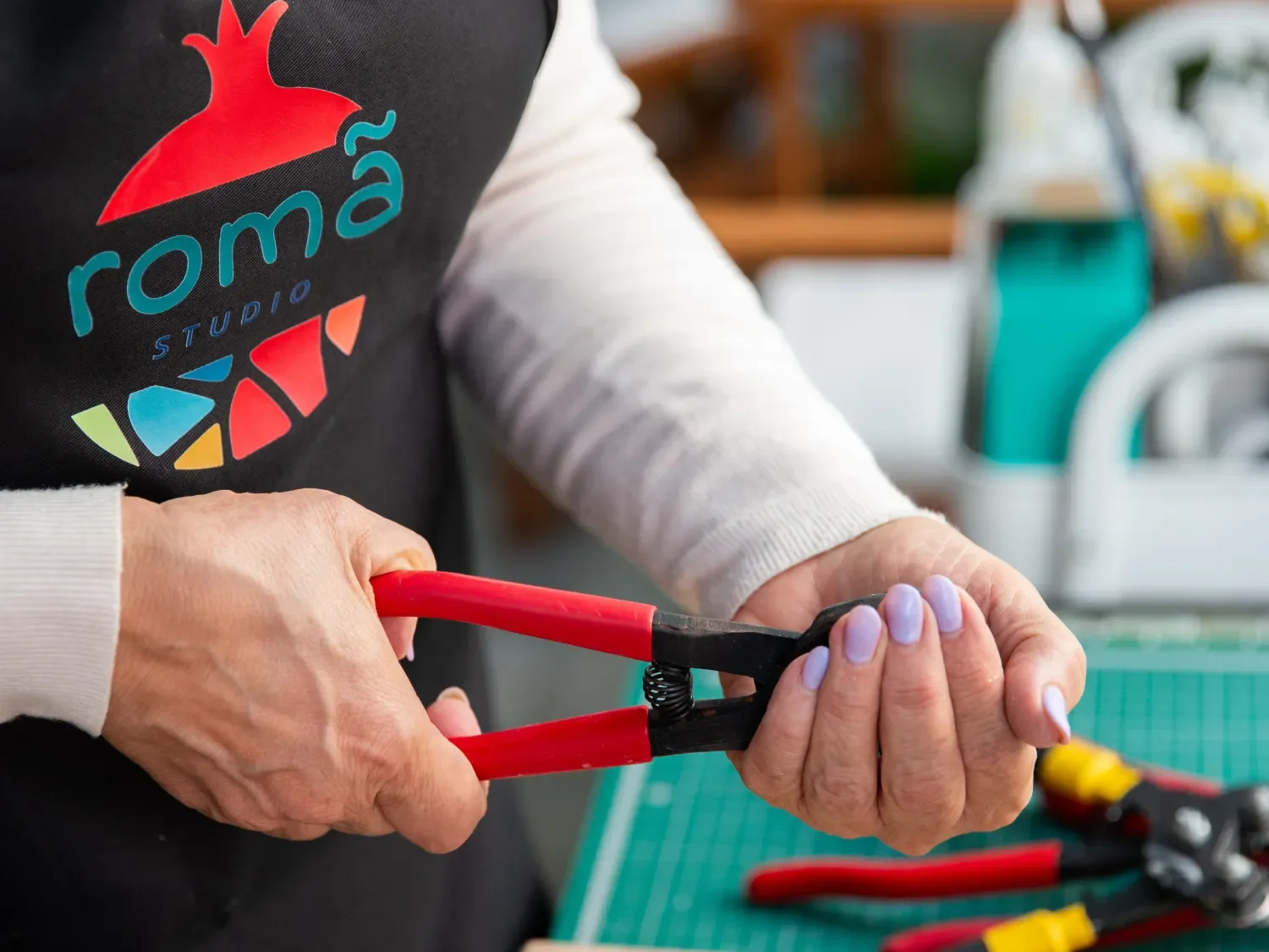 A person holding and using a pair of red-handled pliers, wearing a black apron with colorful logo, in a workshop setting.