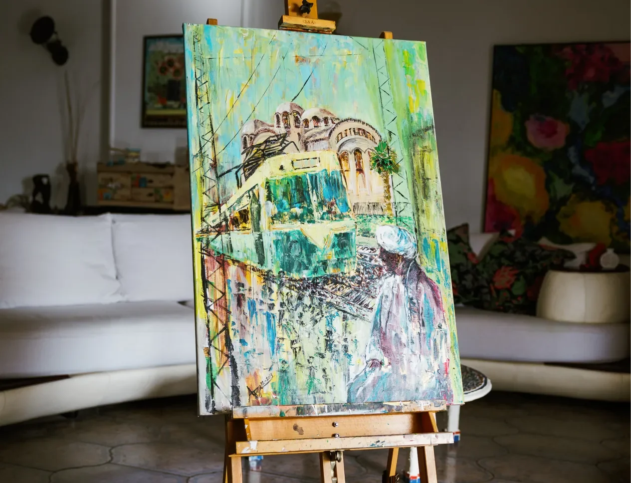 A colorful abstract painting on an easel depicts a person in a white hat and a cityscape with buildings and a green tram, with background paintings and furniture in a living room.