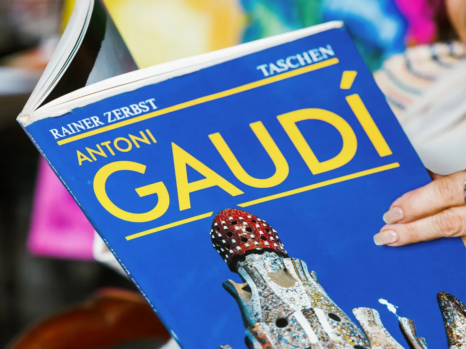 Close-up of a book titled 'Antoni Gaudí' by Rainer Zerbst with a bright blue cover, featuring an image of a mosaic art piece by Gaudí and a person's hand holding the book.