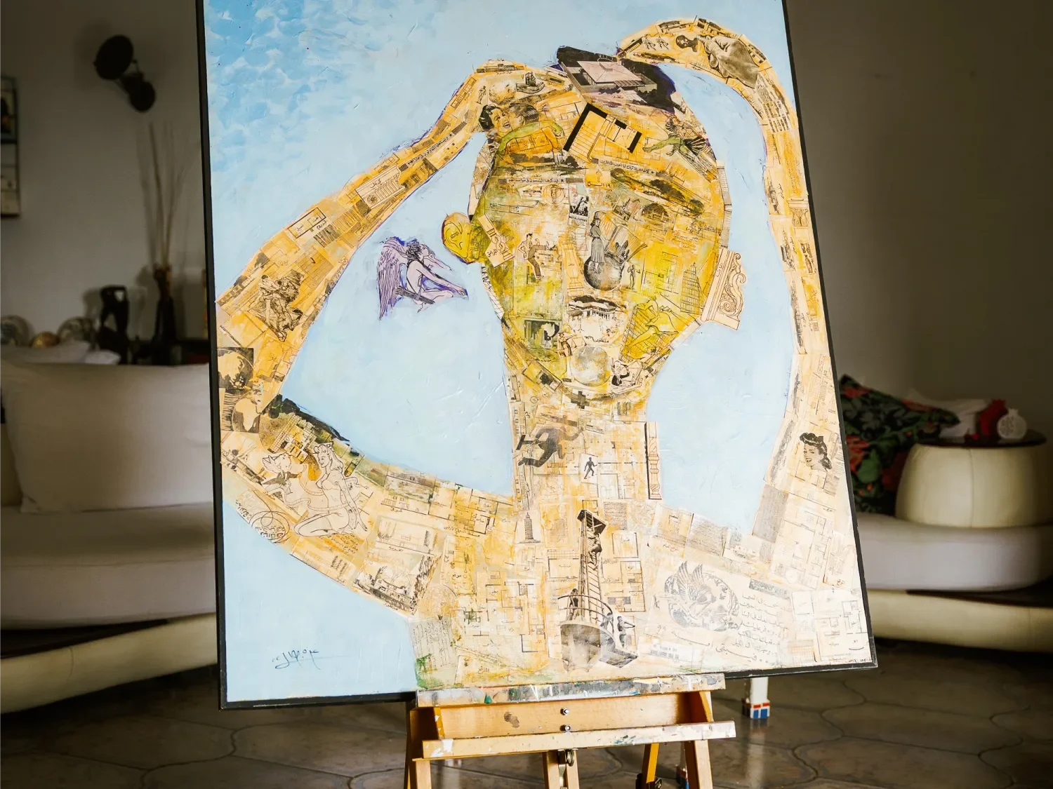 A painting of a person with a yellow head, using a thoughtful pose with hand on forehead, composed of various cutout images and sketches, on an easel in a living room.