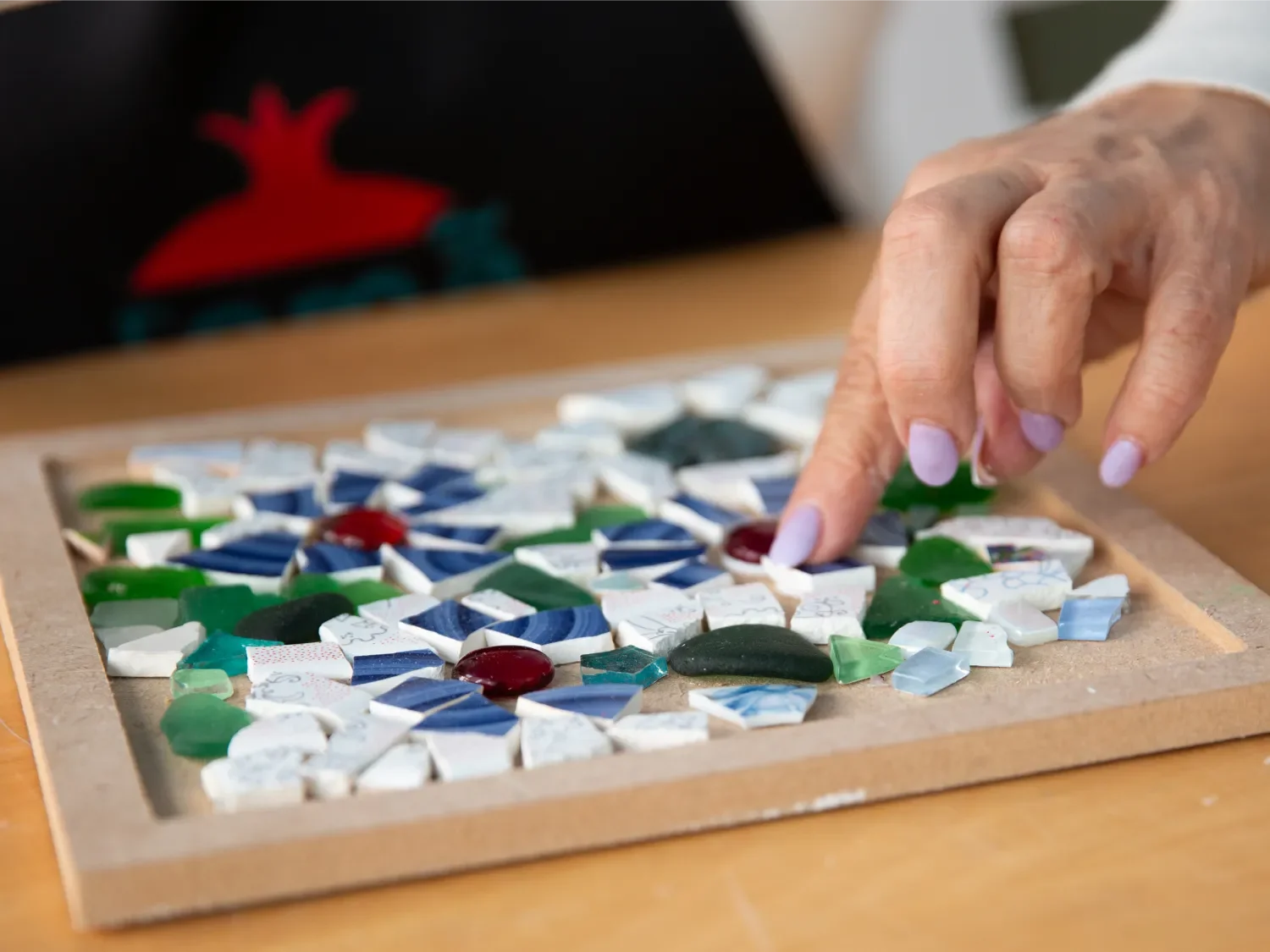 Close-up of a hand arranging pieces of a mosaic on a wooden frame, with various colored and patterned shattered tiles.