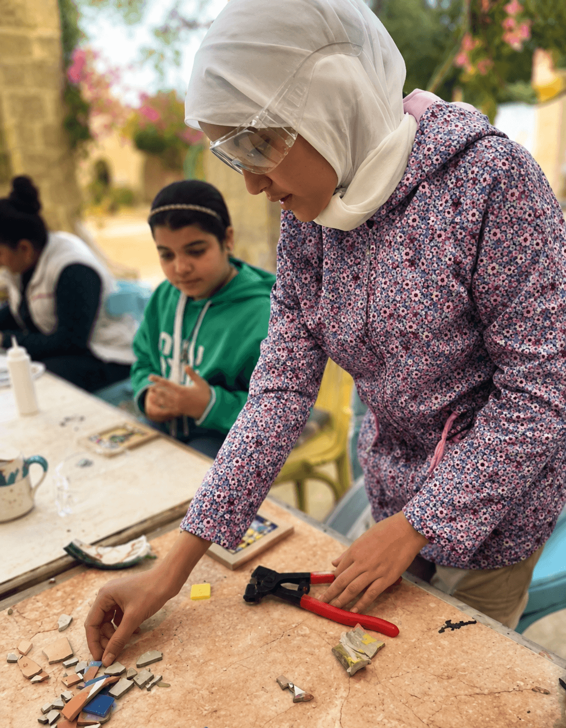 A girl in safety goggles arranges small reclaimed tiles into a mosaic at an outdoor workshop table, with another child working beside her and a garden in bloom behind them.